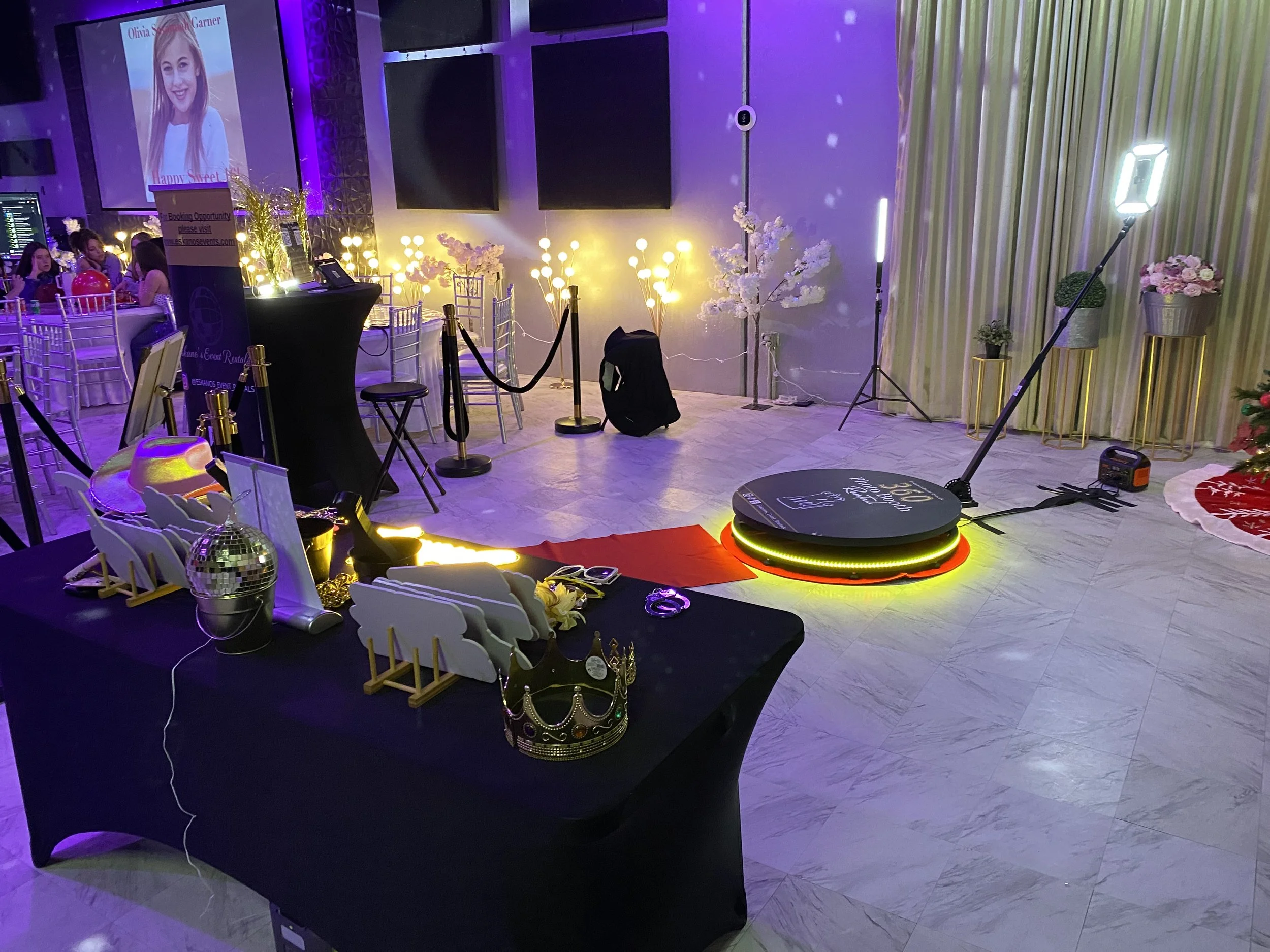 Party room decorated with pink and white flowers, gold accents, and purple lighting. There are table settings with balloons and a large screen displaying a woman's picture and event details.