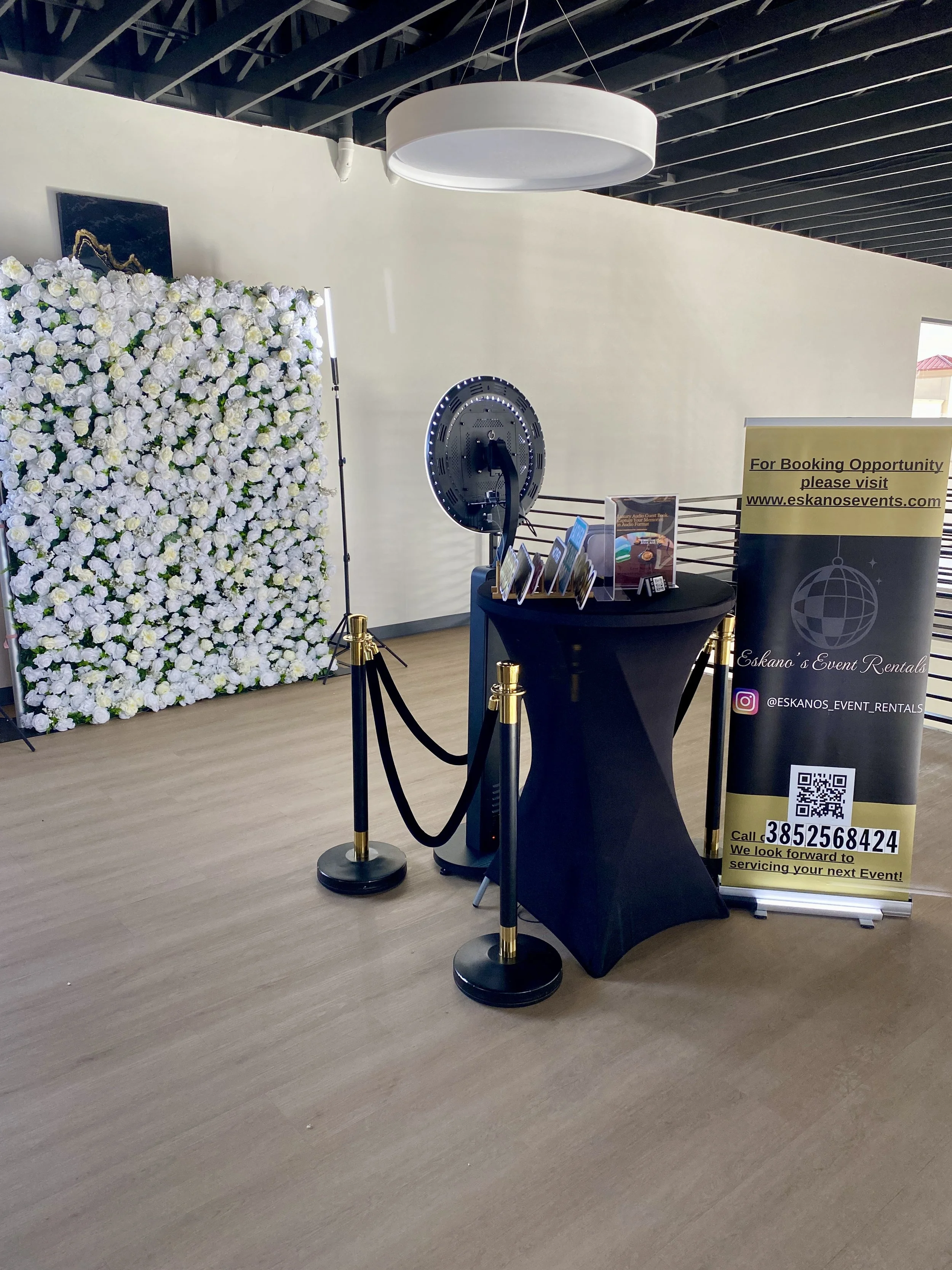 A floral backdrops decorated with white flowers, a photo booth with a spinning wheel, a black table with event flyers, and a banner with contact information for event rentals at Eskano's Event Rentals, inside a room with a wood floor and exposed ceiling beams.