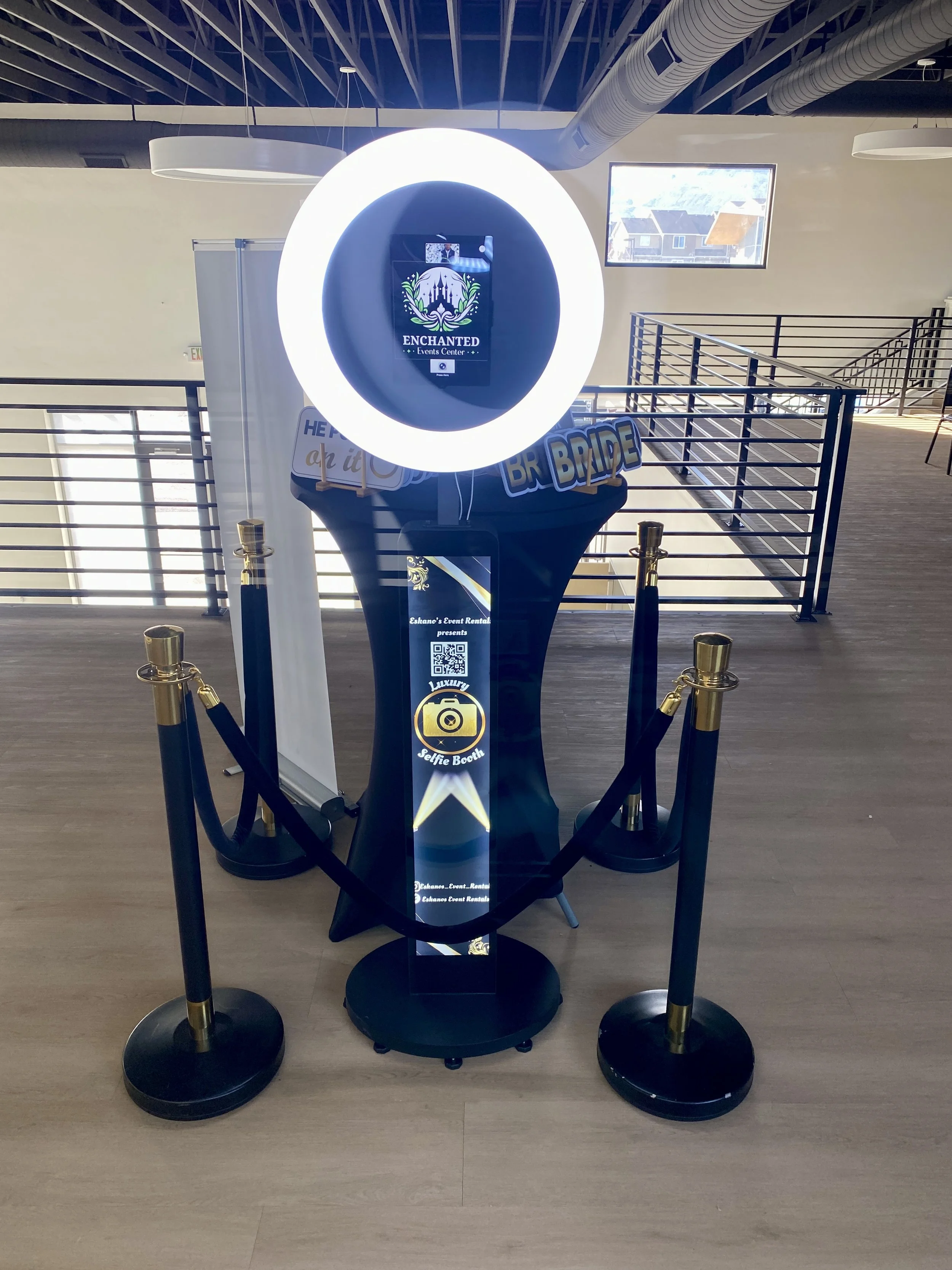 Photo of a self-serve photo booth with a circular ring light, a digital screen, and signs that read 'BRIDE' and 'Selfie Booth'. The booth is enclosed by black ropes with gold stanchions, inside a large, open indoor space with wooden flooring, a few windows, and modern ceiling fixtures.