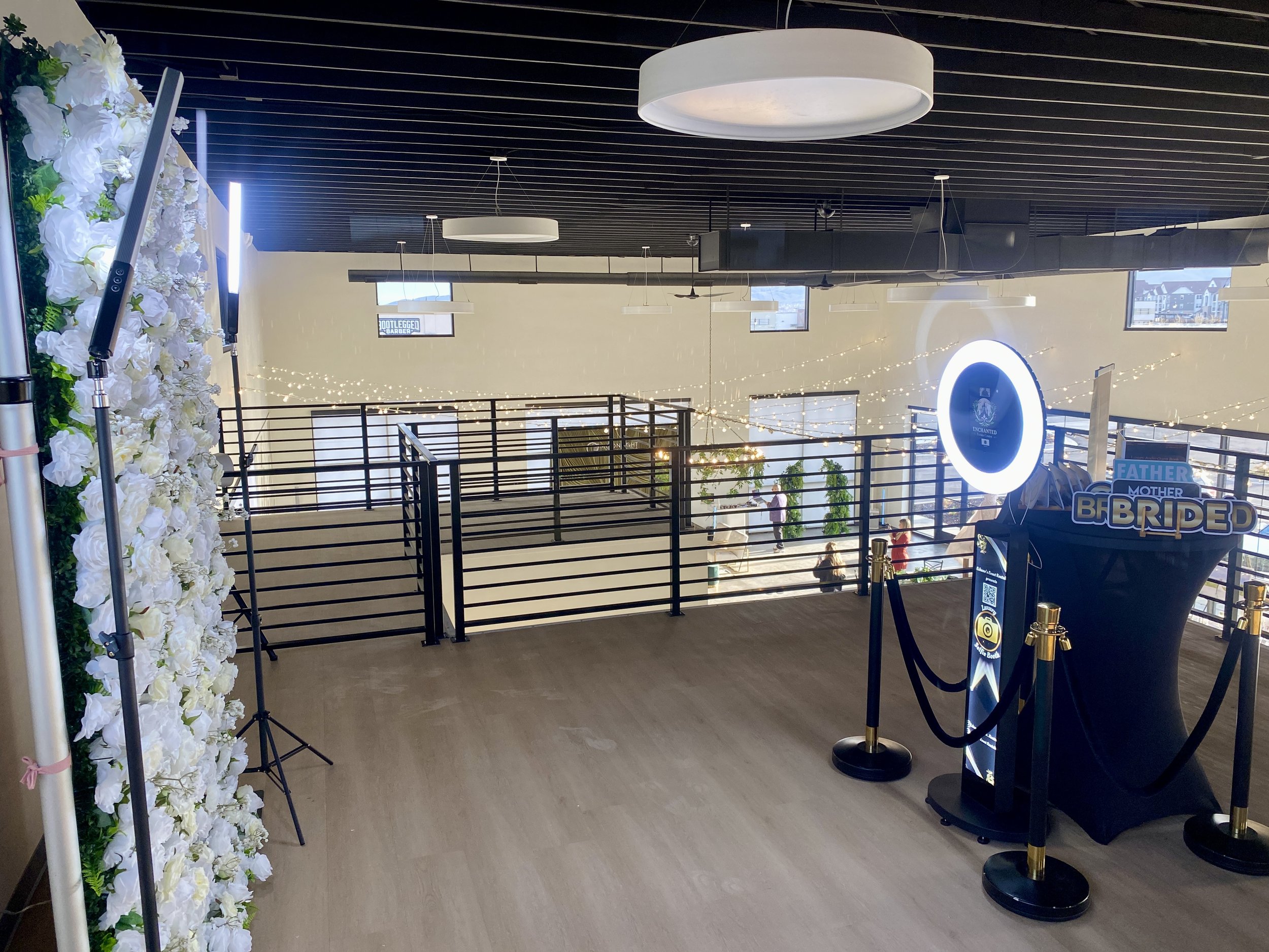 Indoor event space decorated with white flowers on the left, string lights, and a sign that says "Bride" near a black stanchion with a gold rope. There are large circular ceiling lights, windows with views of a cityscape, and a balcony with black railings.