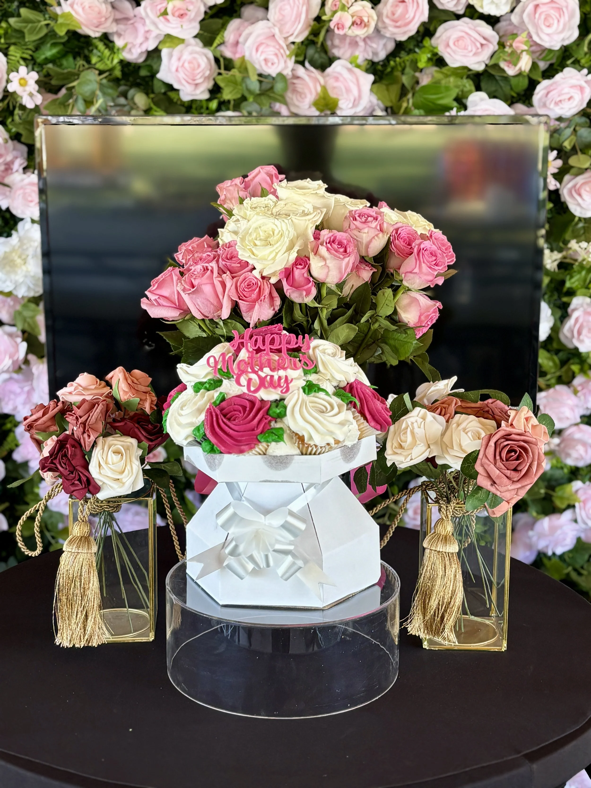 A floral arrangement of pink and white roses on a black table, with a small cake decorated with pink and white roses, a 'Happy Mothers Day' sign, and golden tassels, surrounded by greenery and pink roses in the background.