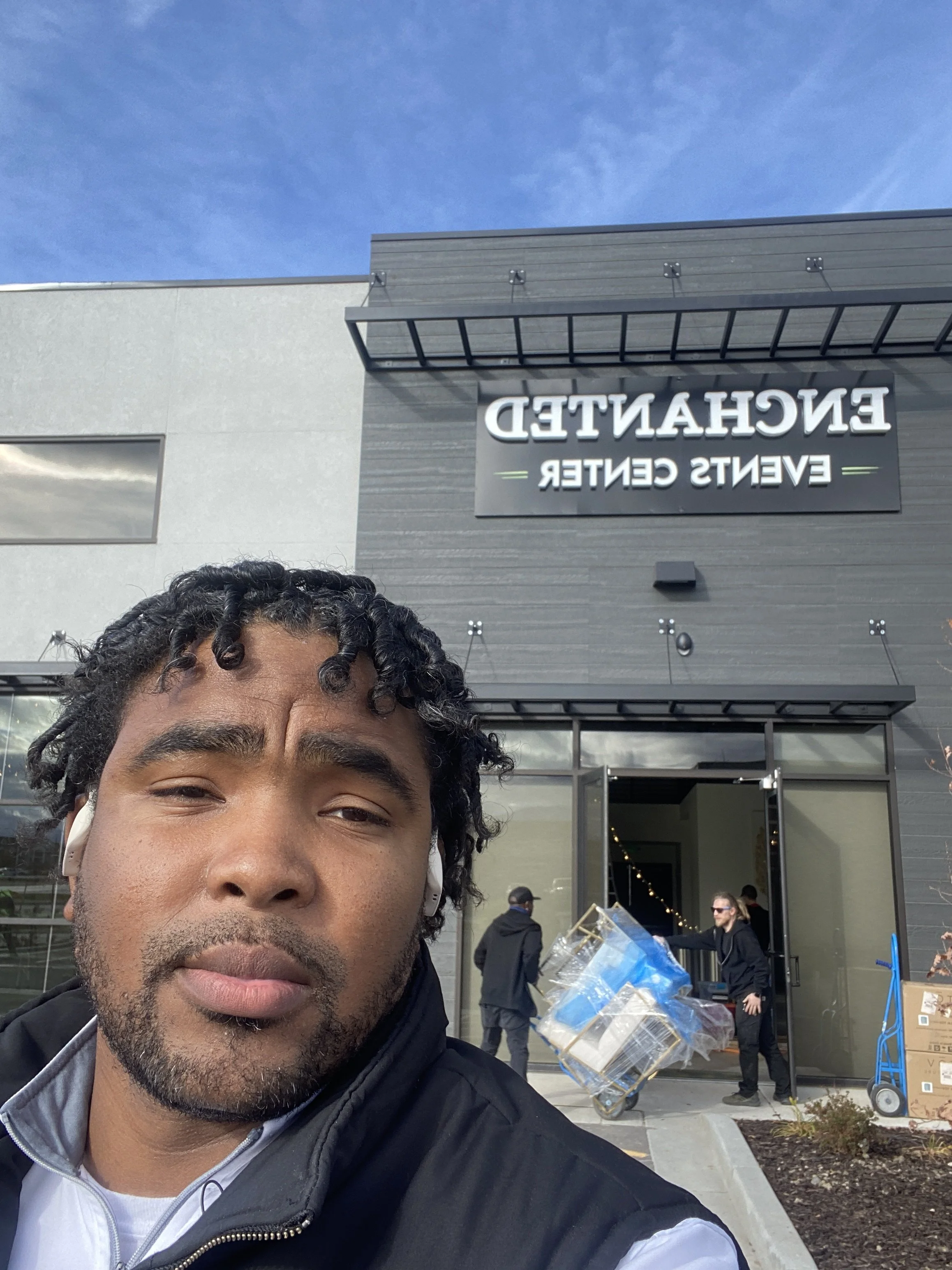 A man with curly hair and a beard taking a selfie outside a modern building with a sign that reads 'ENCHANTED EVENTS CENTER.' There are other people and equipment visible near the entrance of the building.