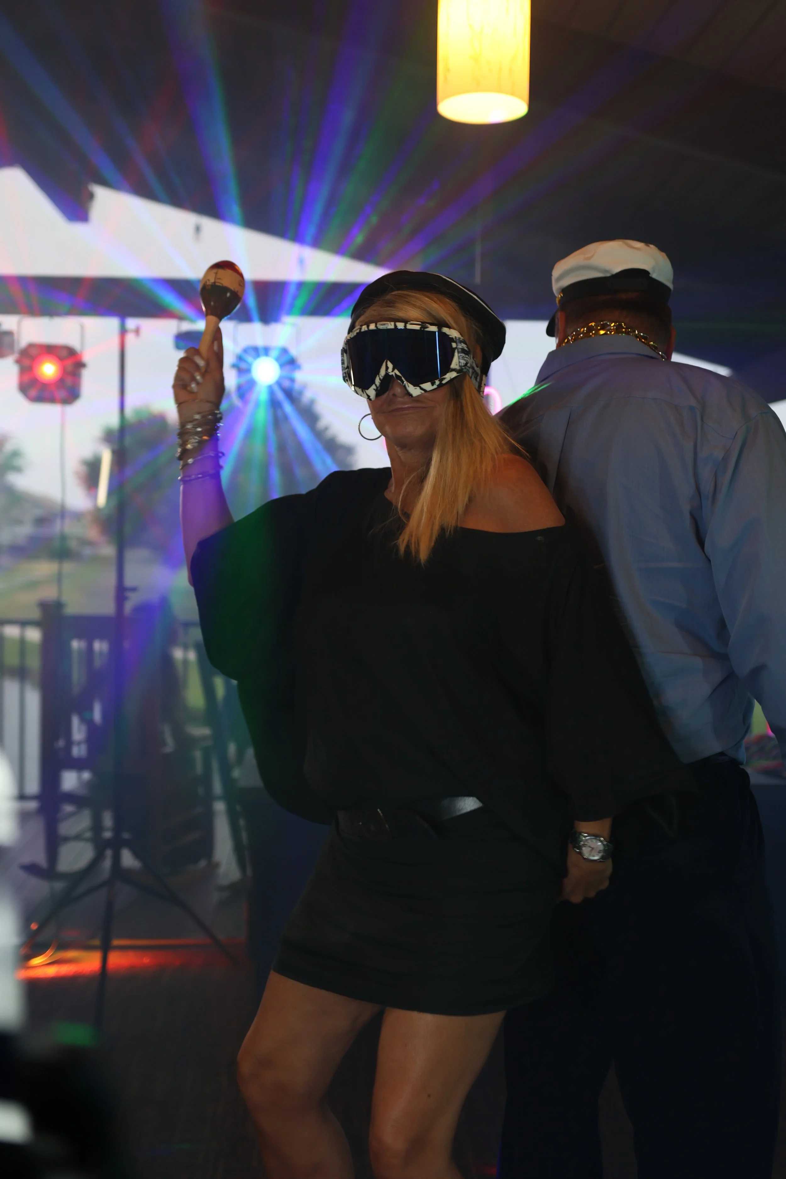 A woman with long, blonde hair wearing large goggles, a black off-shoulder top, and black shorts, holding a maraca in a setting with colorful laser lights and a disco ball, possibly at a party or nightclub.