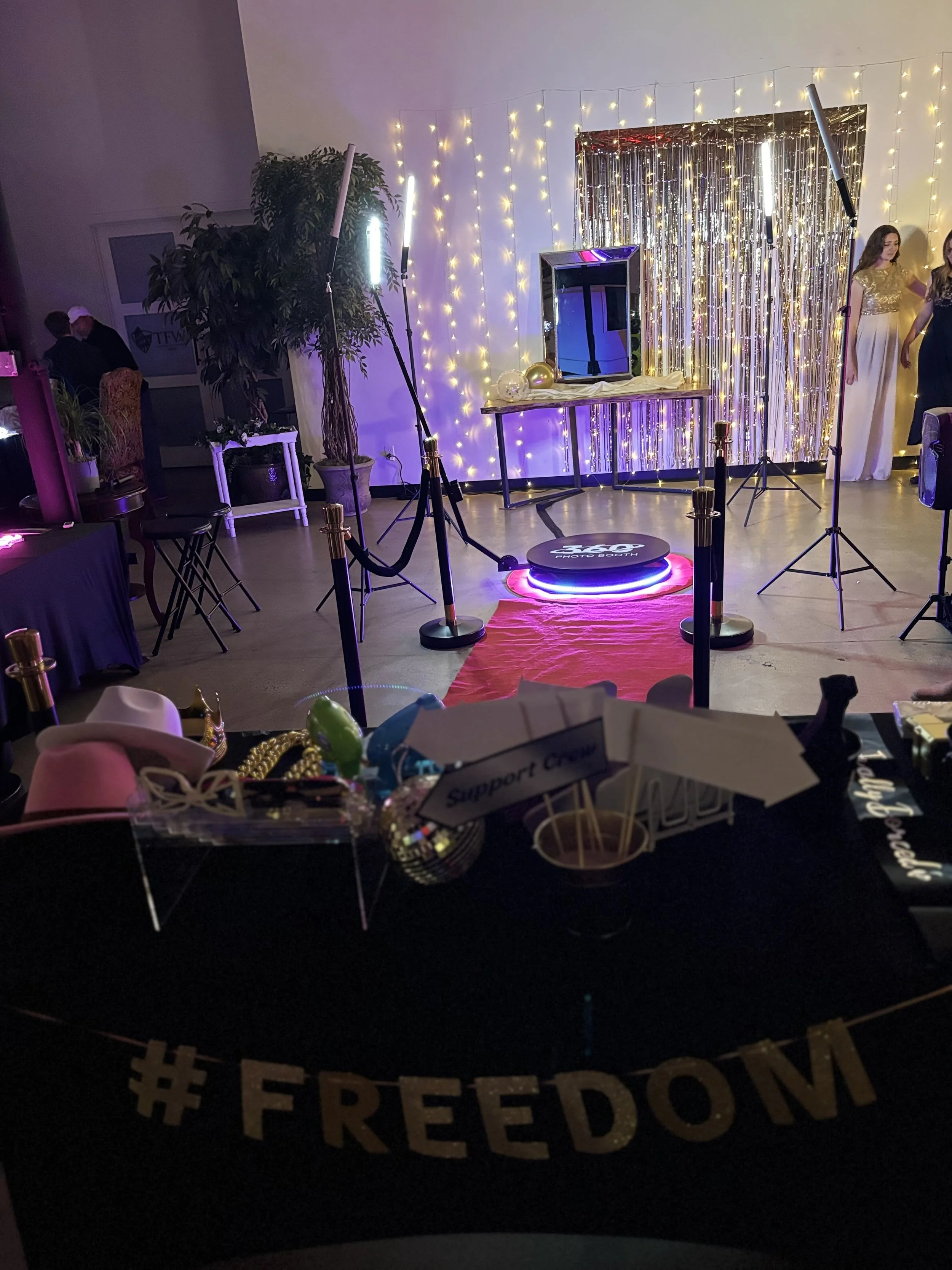 Decorative photo booth area with string lights, balloons, a mirrored frame, a red carpet, and people in the background. The foreground features a hoop with '#FREEDOM' written on it and various props.