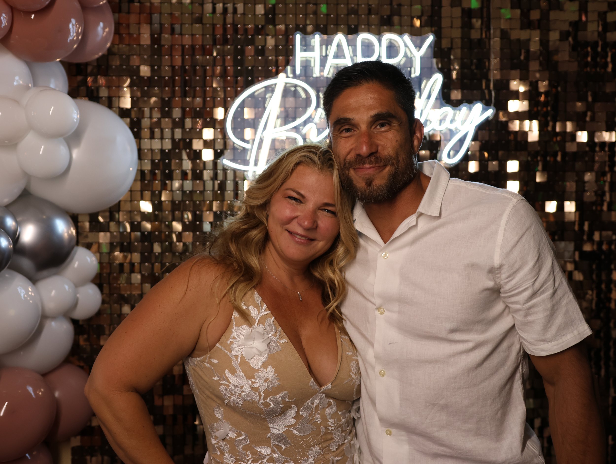A couple at a celebration, smiling and embracing in front of a background with a neon sign that reads 'Happy Birthday' and balloon decorations.