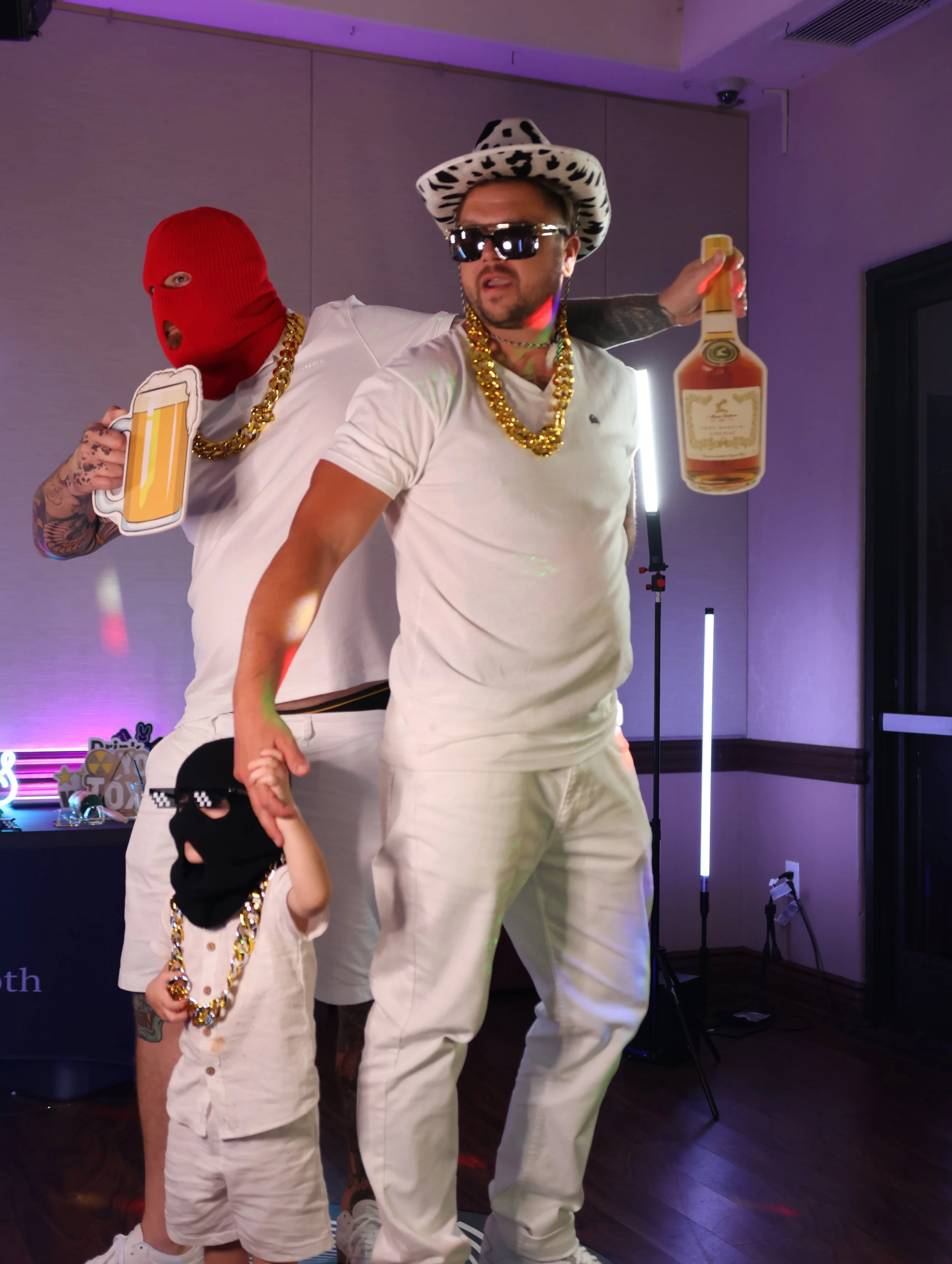 Two men and a child posing with playful expressions, all wearing white clothing and gold chains. One man has dark hair, sunglasses, a black and white animal print hat, and facial hair. The child has a red ski mask, tattoos on the arm, and is holding 