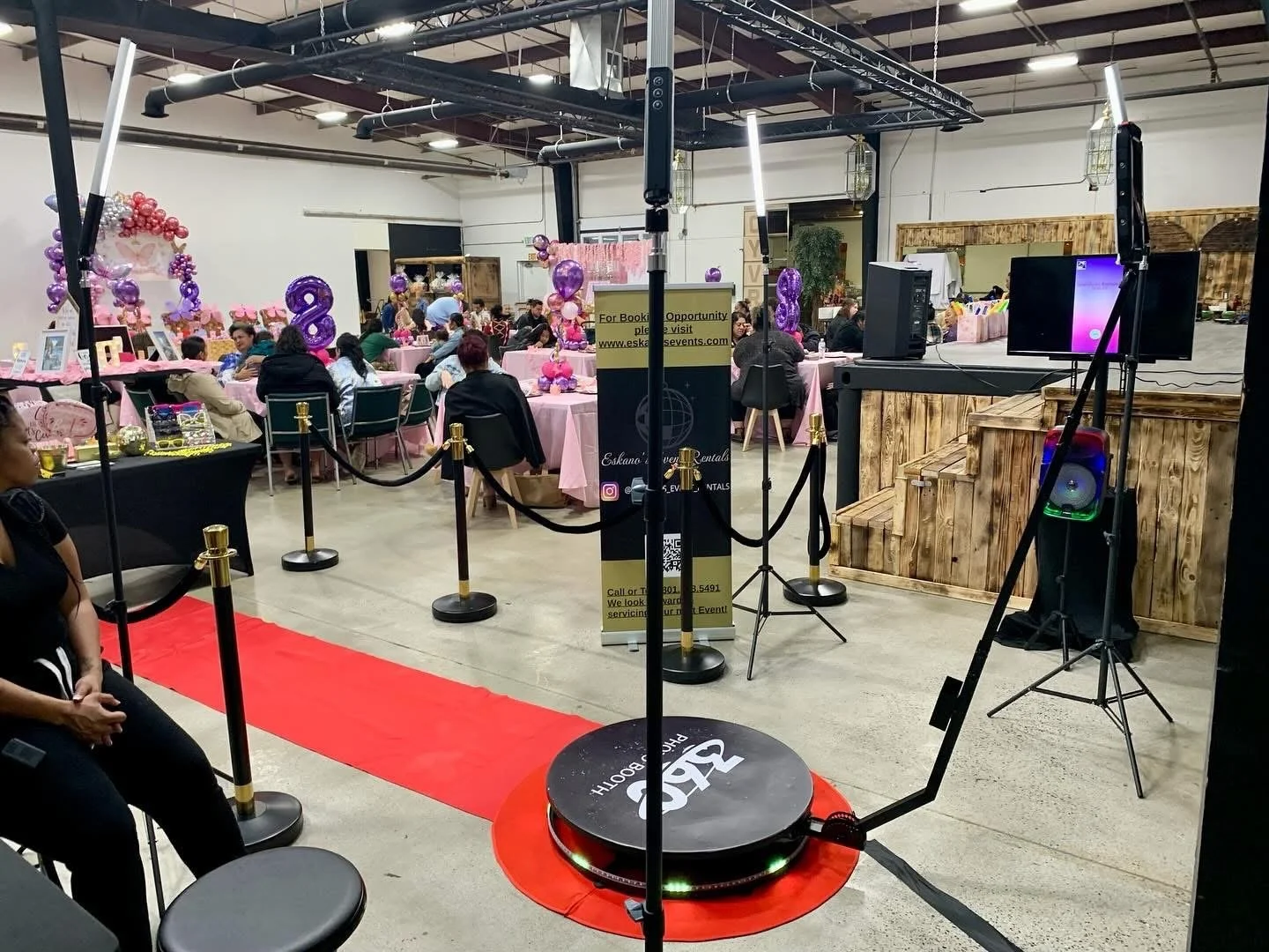 Event space decorated with pink and purple balloons and balloon numbers, set up for a celebration with tables, chairs, and a small stage, with a robot serving as a DJ in the foreground.