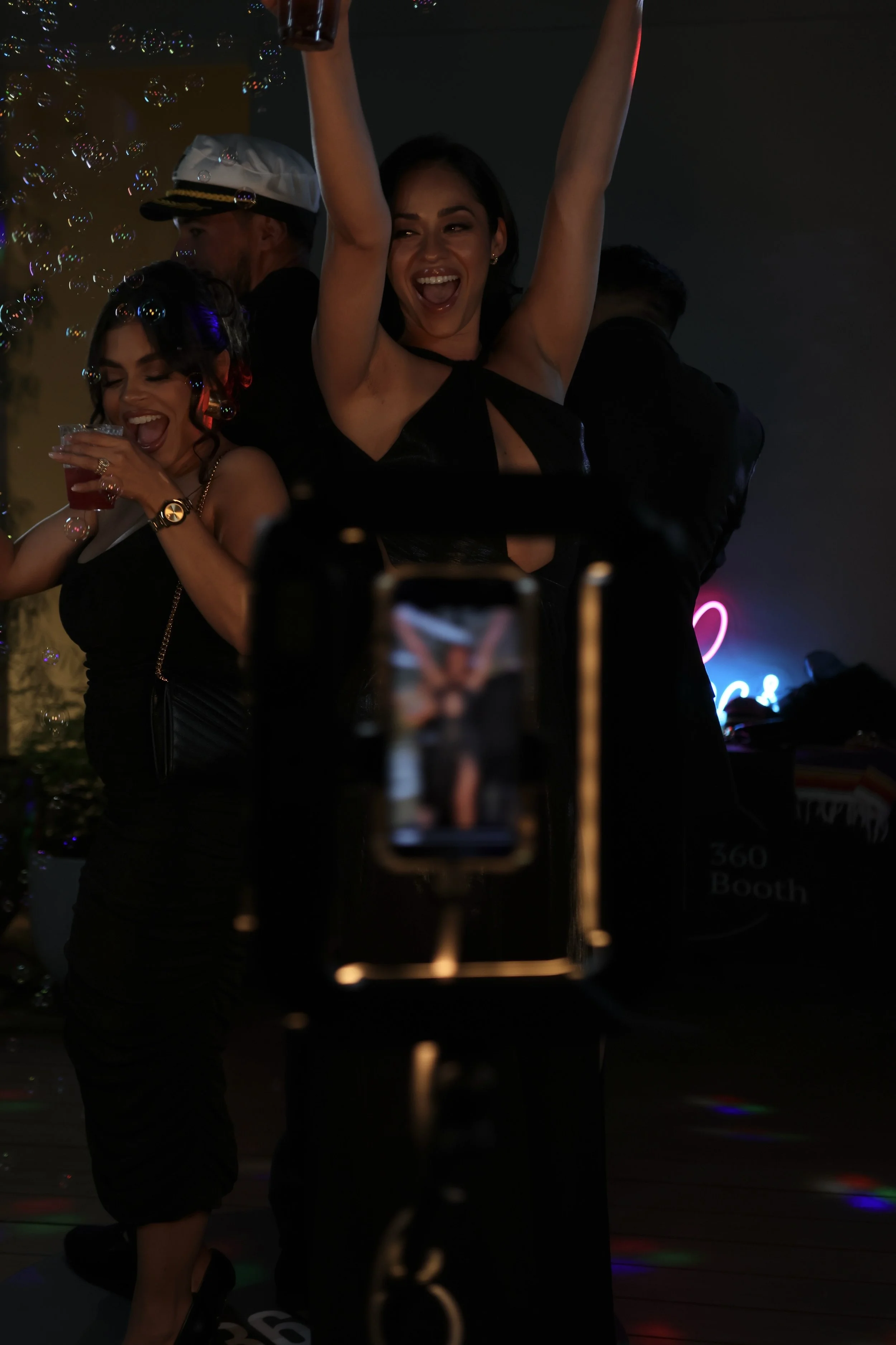 Two women celebrating at a night party, one holding a drink and the other raising her arms in excitement, with bubbles and colorful lights around.