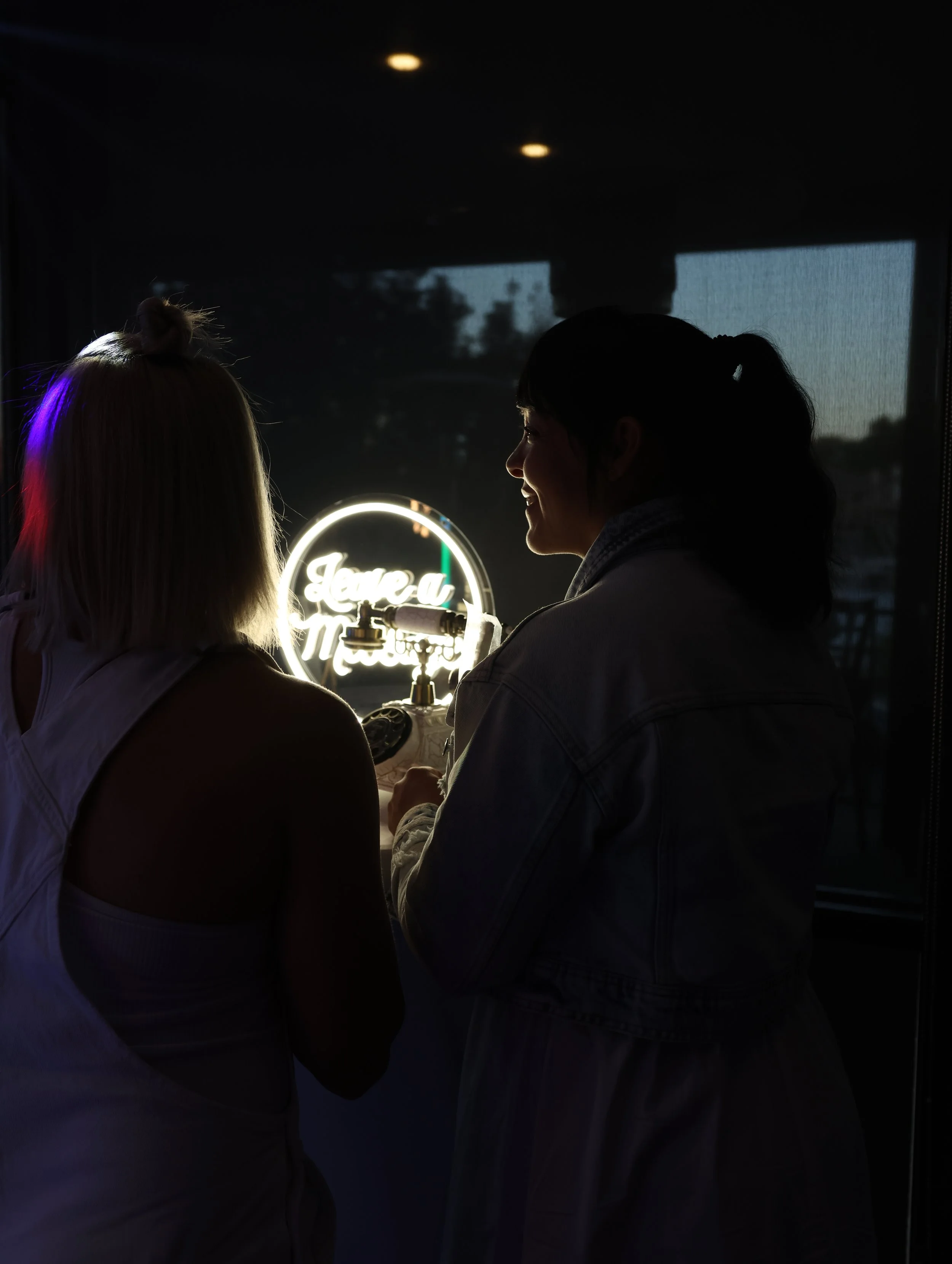 Two women are sitting together during sunset or indoors with dim lighting, smiling and having a conversation, with a neon sign in the background.