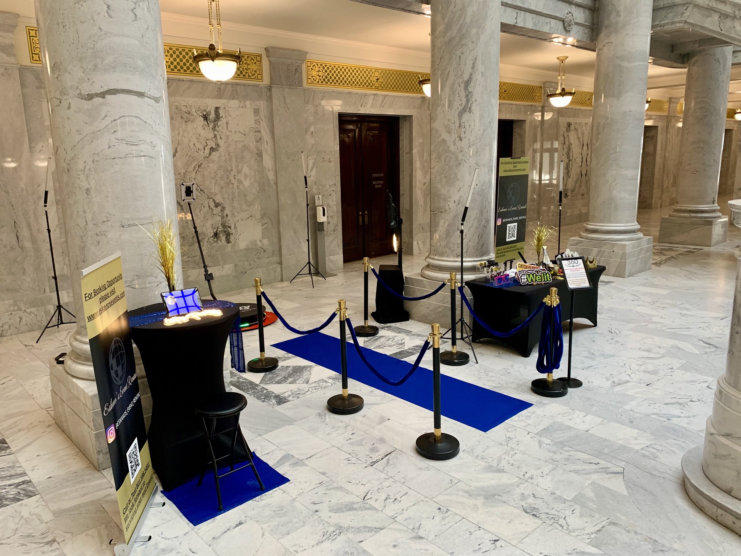 Hotel lobby with marble pillars and flooring, featuring a registration desk, hanging lights, and signs for event rentals and booking opportunities. A blue carpet runs through the area enclosed by stanchions with gold accents.