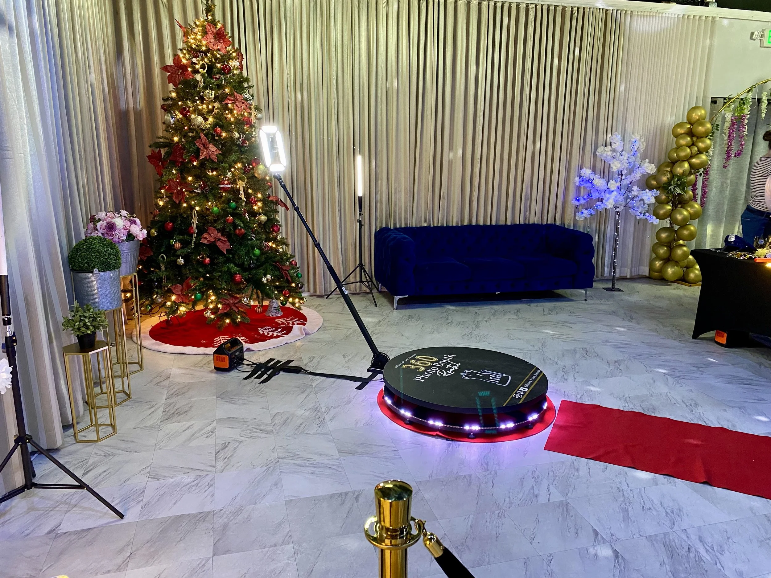 Decorated Christmas tree with ornaments and lights, beside a purple floral arrangement, a red and white Christmas rug, and inflatable golden and white balloons, with lighting equipment and a blue sofa in the background.