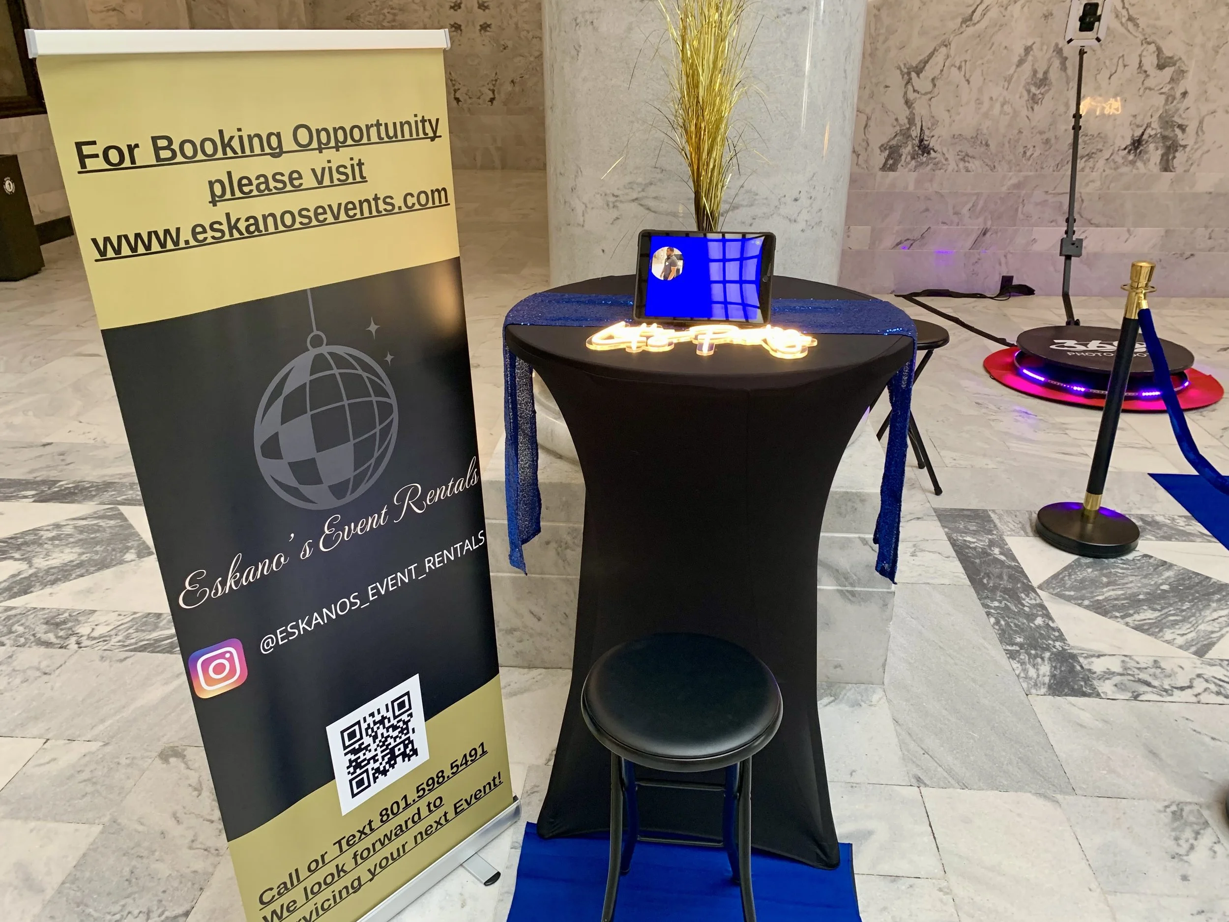 A yellow sign on a stand promoting event bookings at Eskano's Event Rentals with a QR code, a small round table with a black seat and a decorative plant in a marble vase behind it, and a photo booth with a round black base and blue LED lights, set up in a marble-finished lobby area.
