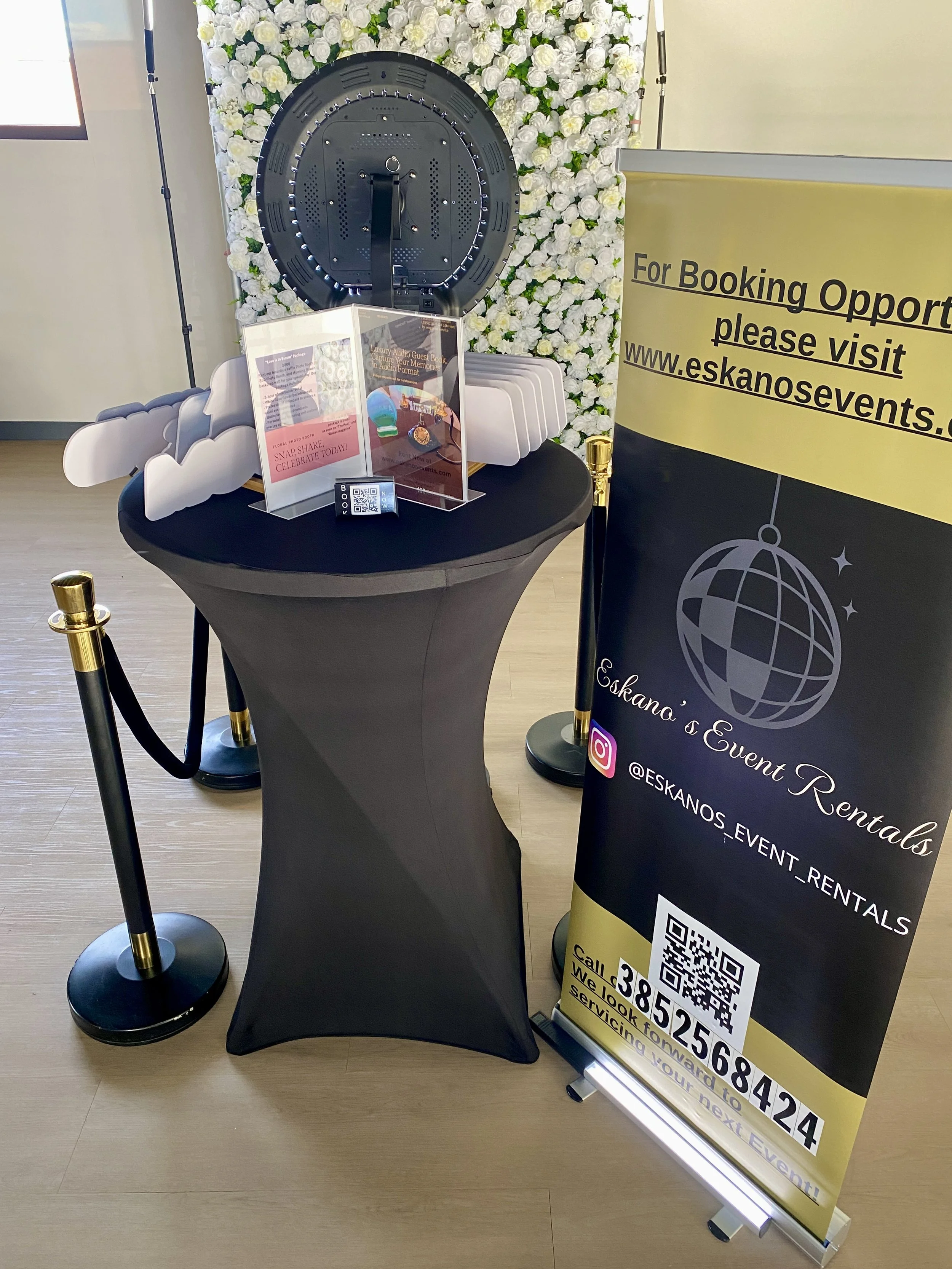A promotional display for Eskano's event rentals, featuring a black table, a yellow and black sign with booking information, and a floral backdrop with white roses. There is also a photo booth sign with a QR code and a small transparent box for guest