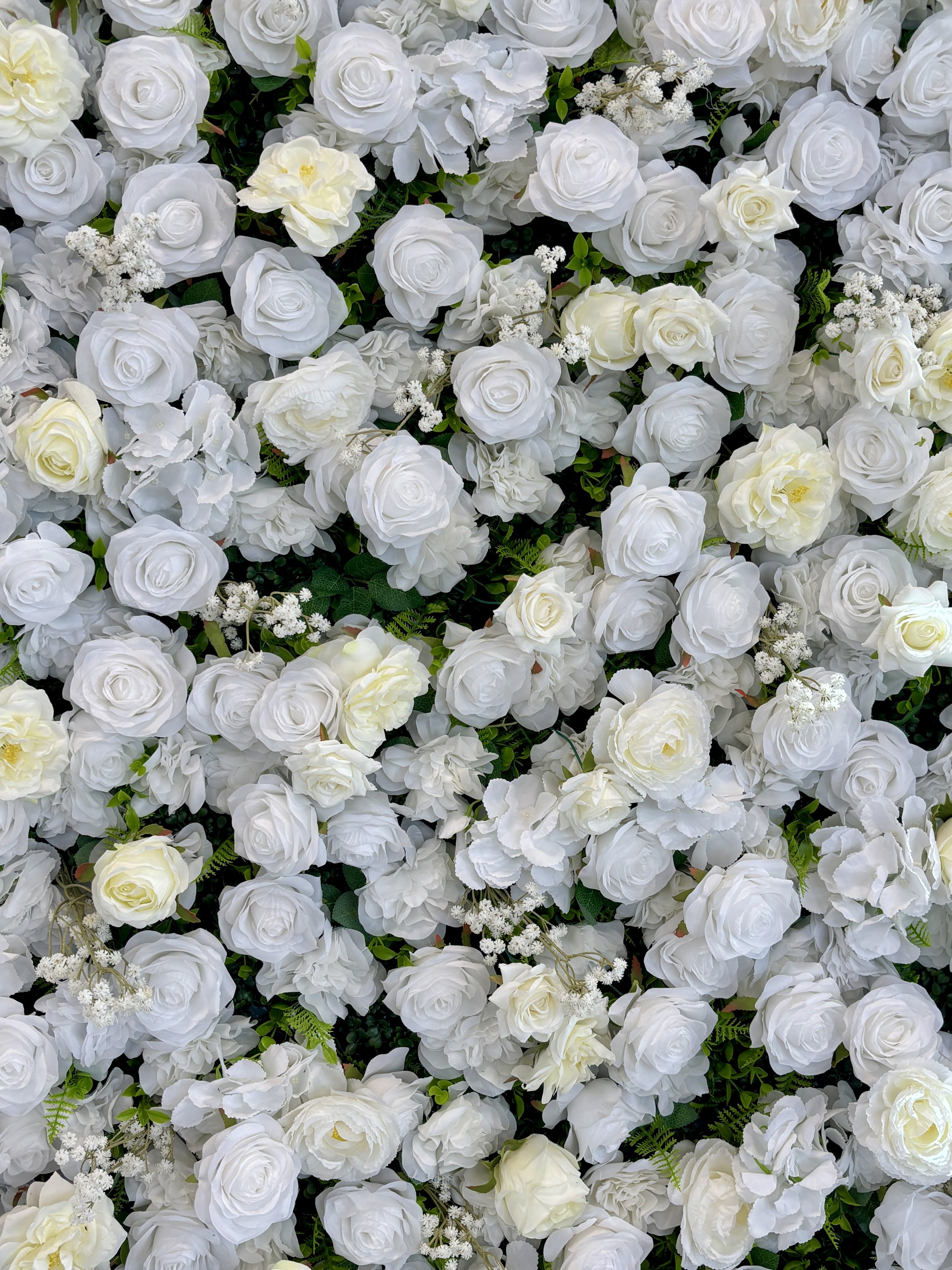 A close-up view of a wall covered in various white flowers, including roses, hydrangeas, and small filler flowers, accompanied by green foliage.