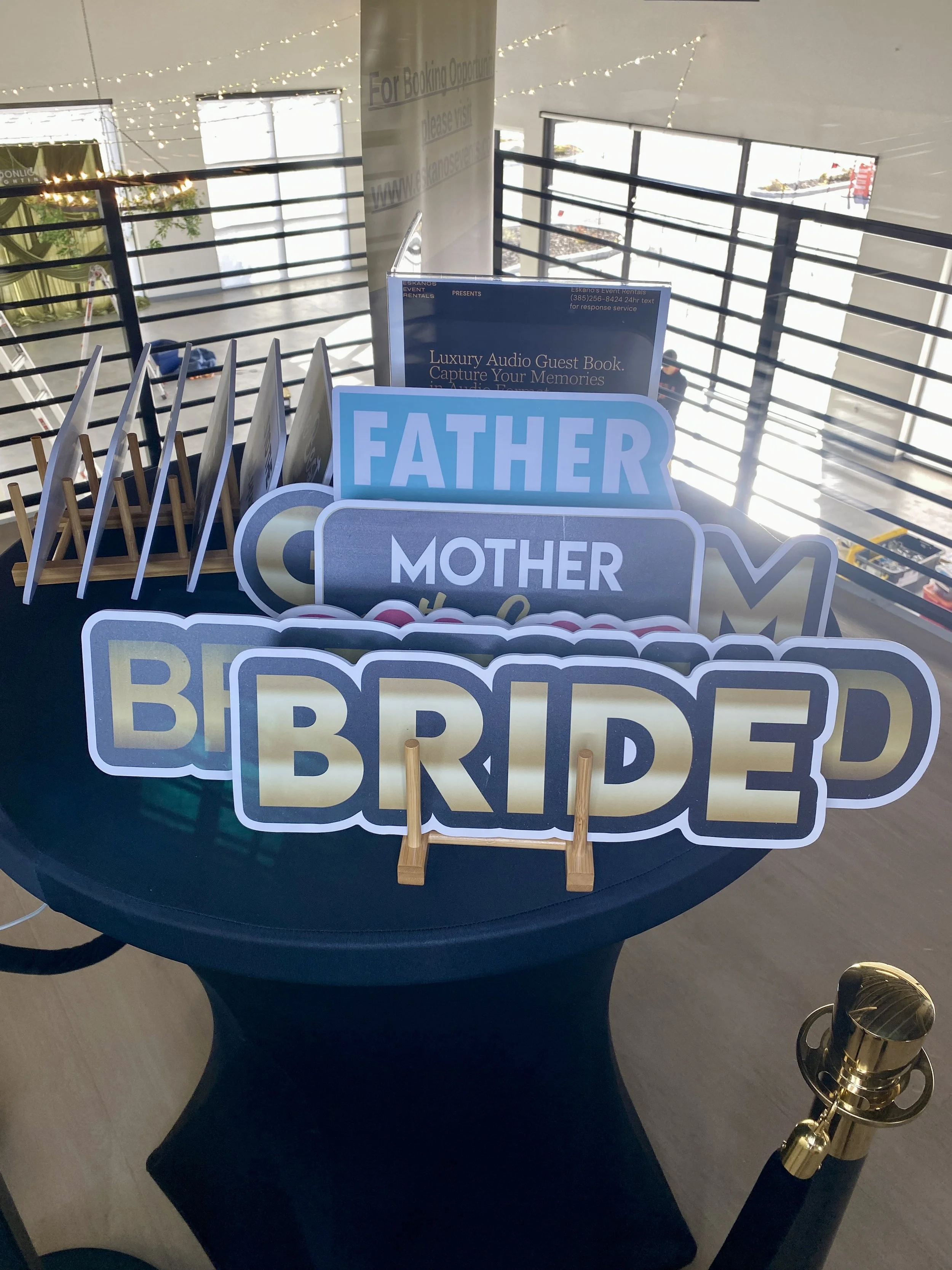 Display of photo booth props including signs that say 'Father', 'Mother', and 'B ride' on a table with a blue cover. In the background, there are window and string lights, with a partial view of a sign and some decorations.