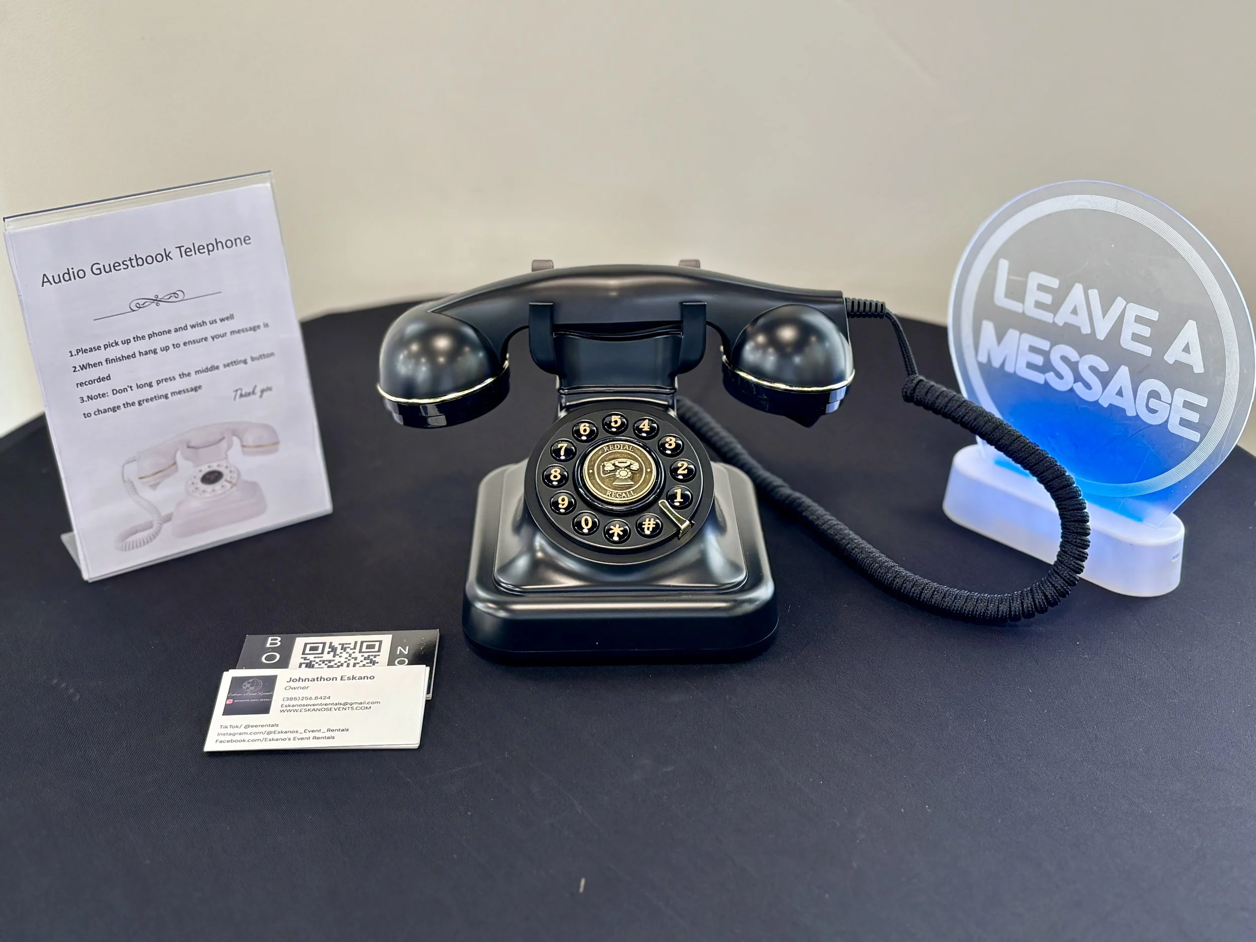 A vintage black rotary telephone with a circular dial and a corded handset, placed on a black surface. To the right, a illuminated sign reads 'LEAVE A MESSAGE' in white text. To the left, a clear plastic stand displays a printed sheet titled 'Audio G