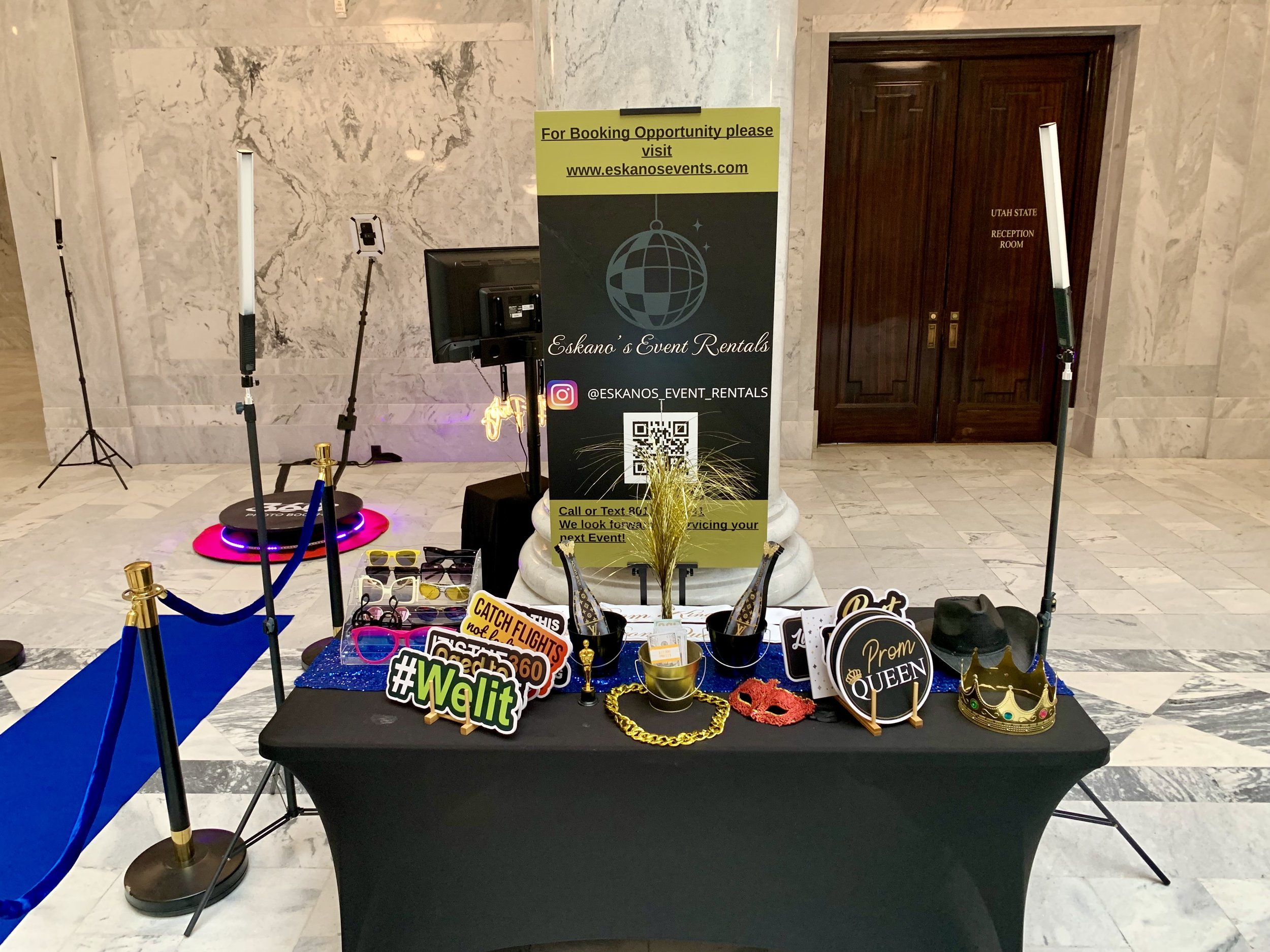 Display table with various photo booth props including crowns, glasses, signs, and accessories, at an event venue entrance with a sign for Eskano's Event Rentals and a sign for booking through a website.