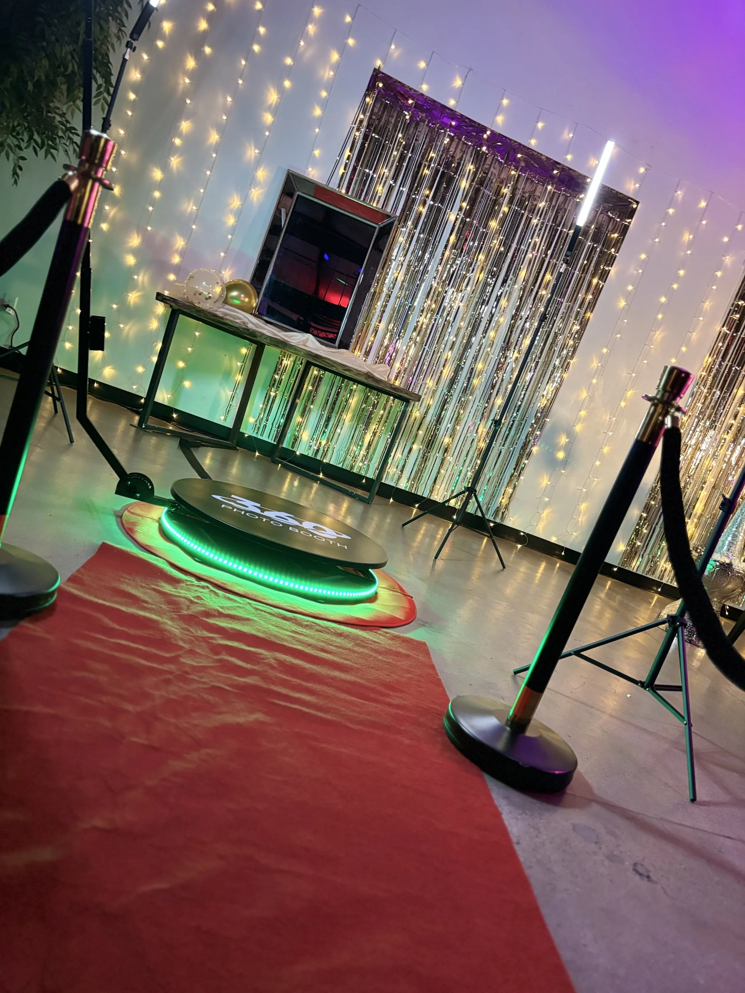 Photo of a photo booth setup with a backdrop of twinkling string lights, metallic streamers, and a table with balloons and decorations in a party or event setting. The photo booth platform has an illuminated sign reading 'PHOTO BOOTH.'