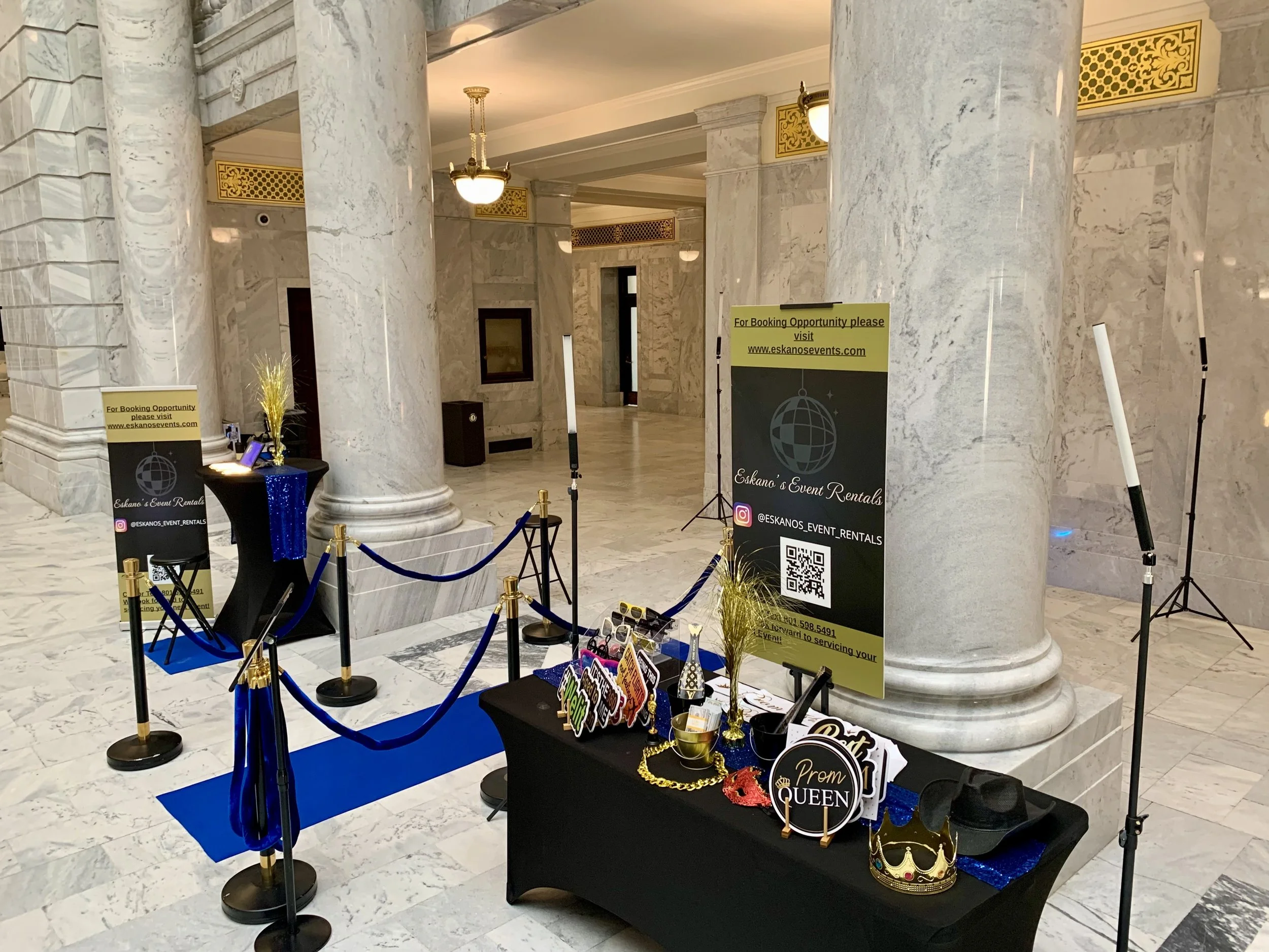 Indoor event space with marble columns and a table decorated for a prom event, including signs and a crown, with banners promoting event rentals and a QR code, set up with velvet ropes and decorative props.