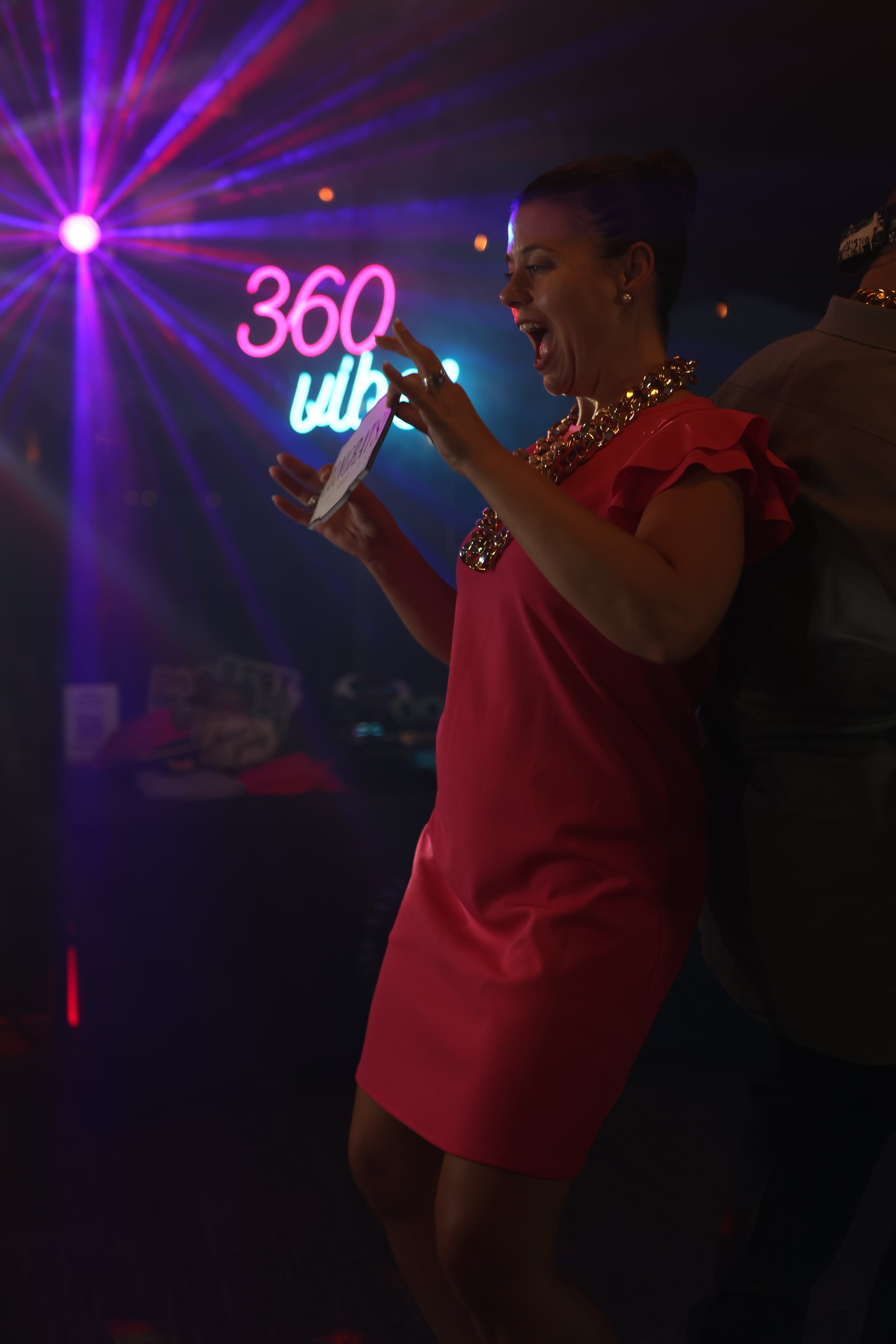 A woman in a red dress with ruffled sleeves and a gold necklace smiling and laughing at a party or nightclub. She is holding a piece of paper or a card and pointing at it. There are colorful neon lights and a sign that reads '360' in the background.