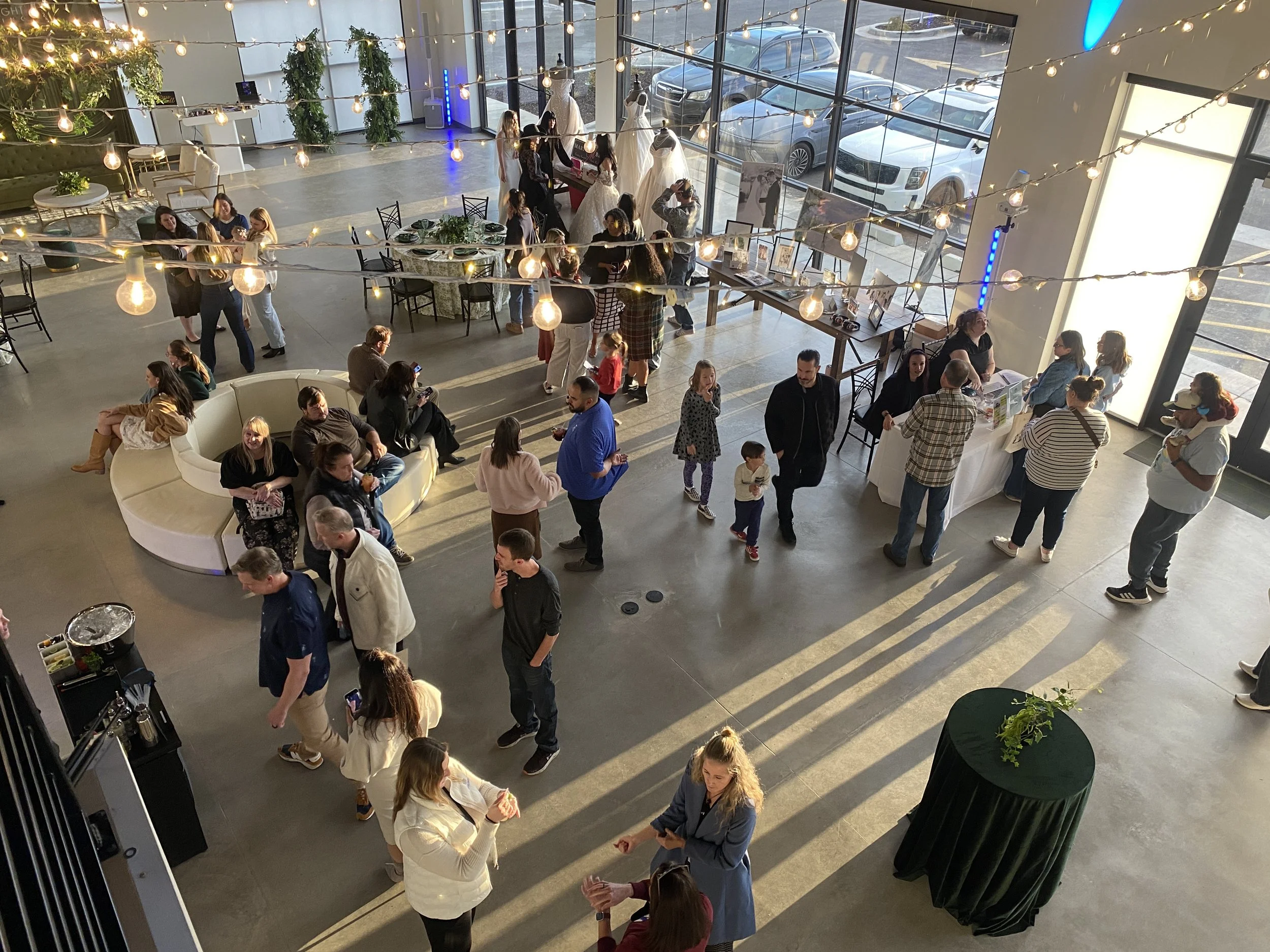 People socializing at a gray interior venue decorated with string lights and a Christmas tree, with a display of wedding dresses on mannequins near large glass windows and several tables and chairs.