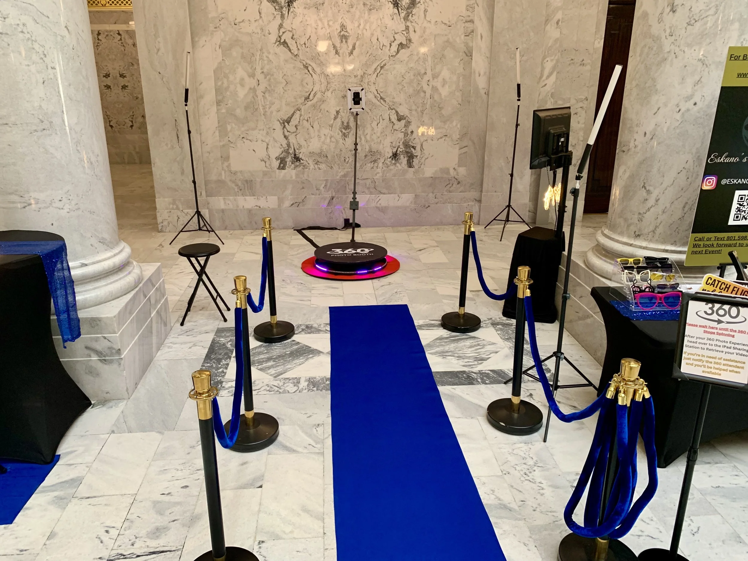 A blue carpet leading to a photo booth with a circular black platform with LED lights, surrounded by black and gold stanchions and blue ropes. The walls are made of white marble with gray veins, and there are lighting equipment and a screen on the right side.