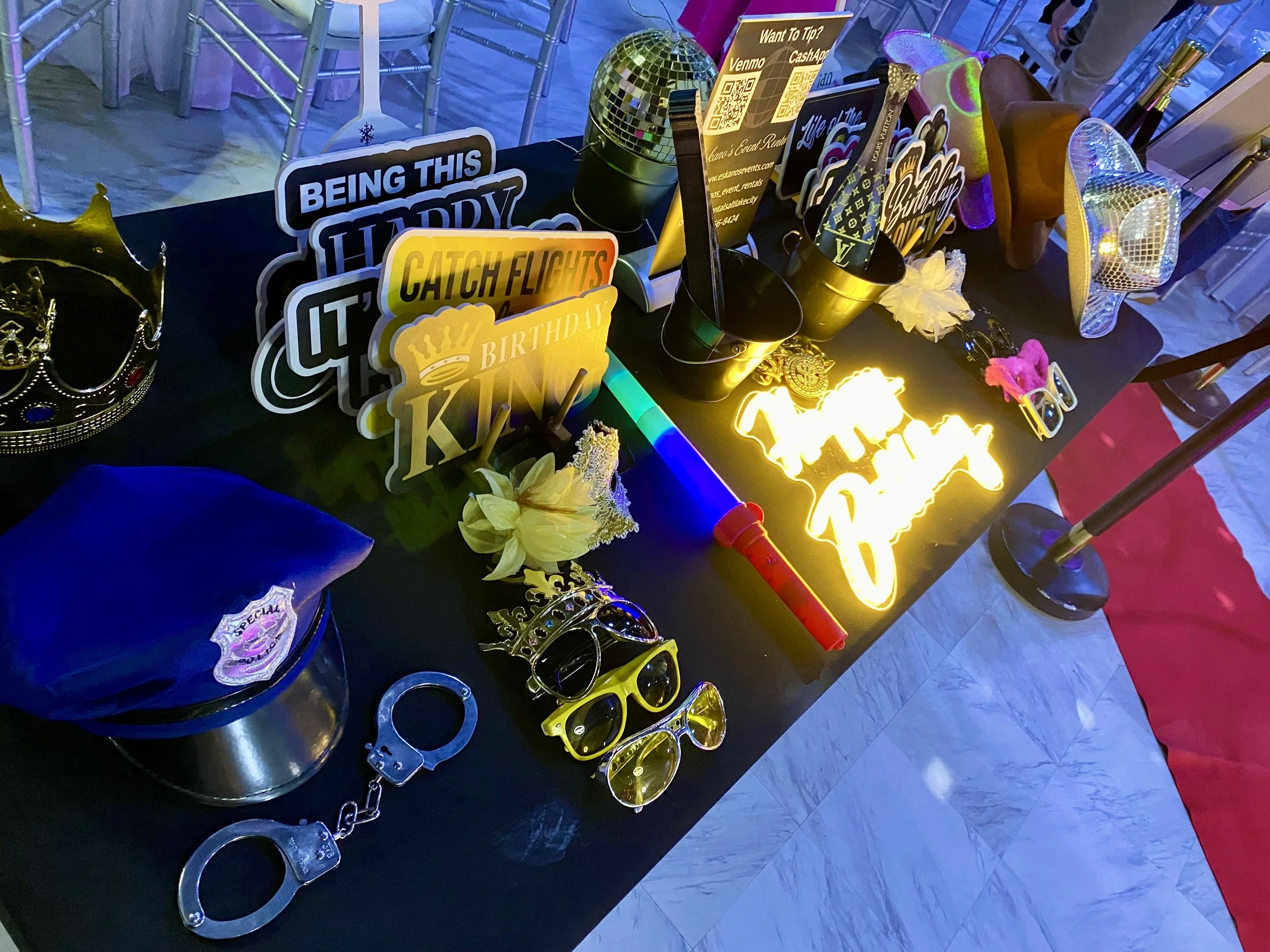 Decorative items for a birthday celebration on a black table, including signs, sunglasses, a disco ball, a crown, and a glowing Texas-shaped sign, with chairs and a pink tablecloth in the background.