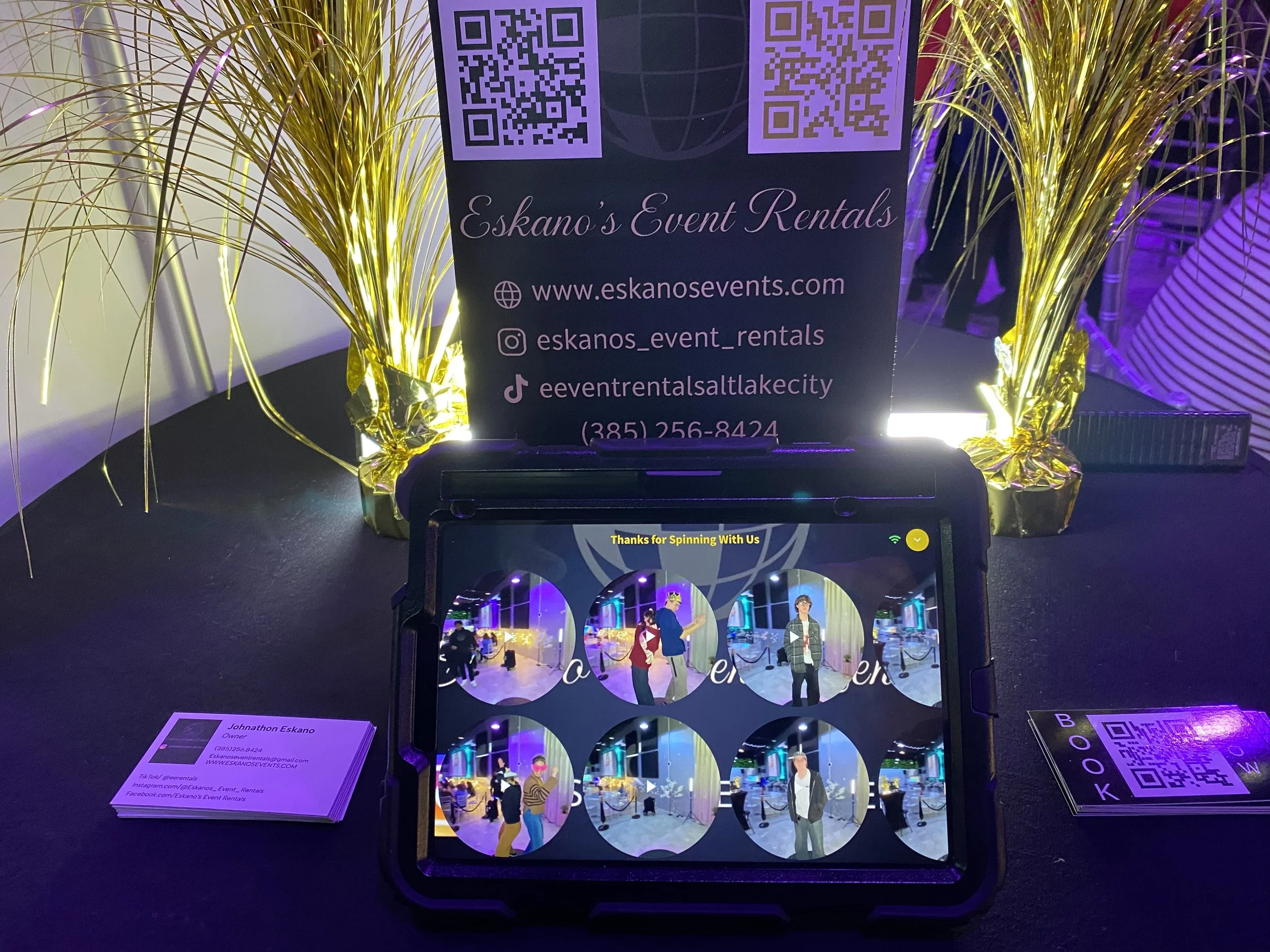 Sign for Eskano's Event Rentals with QR codes, contact information, and decorative gold tinsel around it, set at an event.