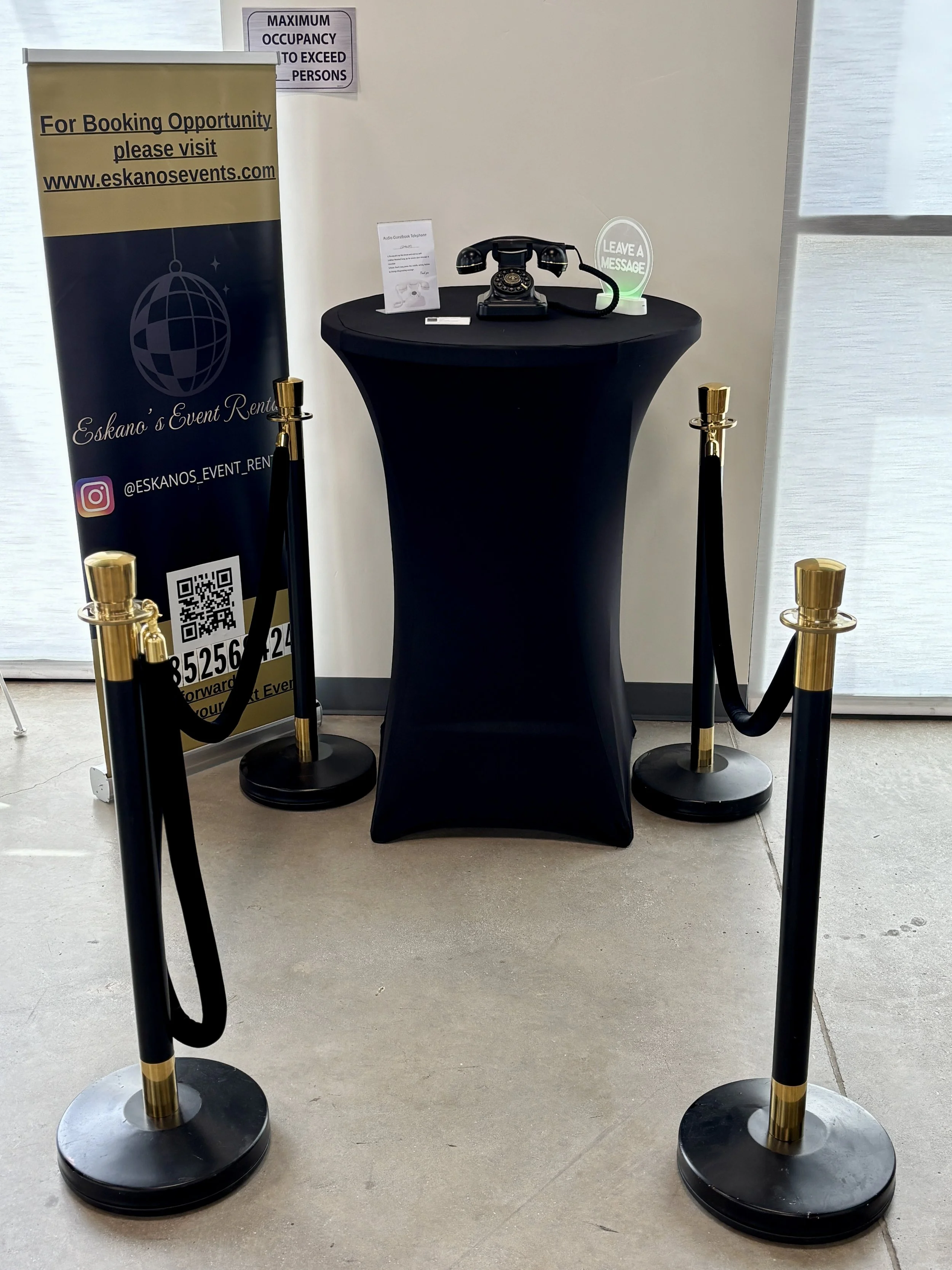 A vintage black rotary telephone on a round black table covered with a black tablecloth. To the right of the phone, there is a small round sign that reads 'Leave a Message'. Behind the table, a standee with a globe design and information for Eskano's