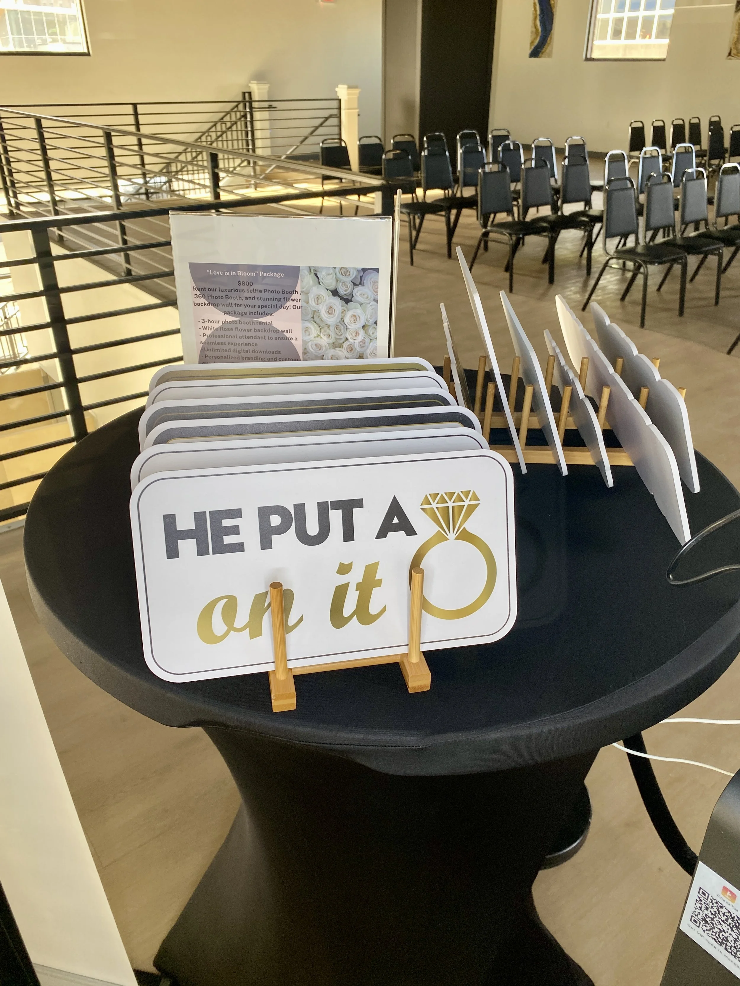 Display table with wedding sign and small white chairs, behind which are empty black chairs, in a room with large windows and beige walls.