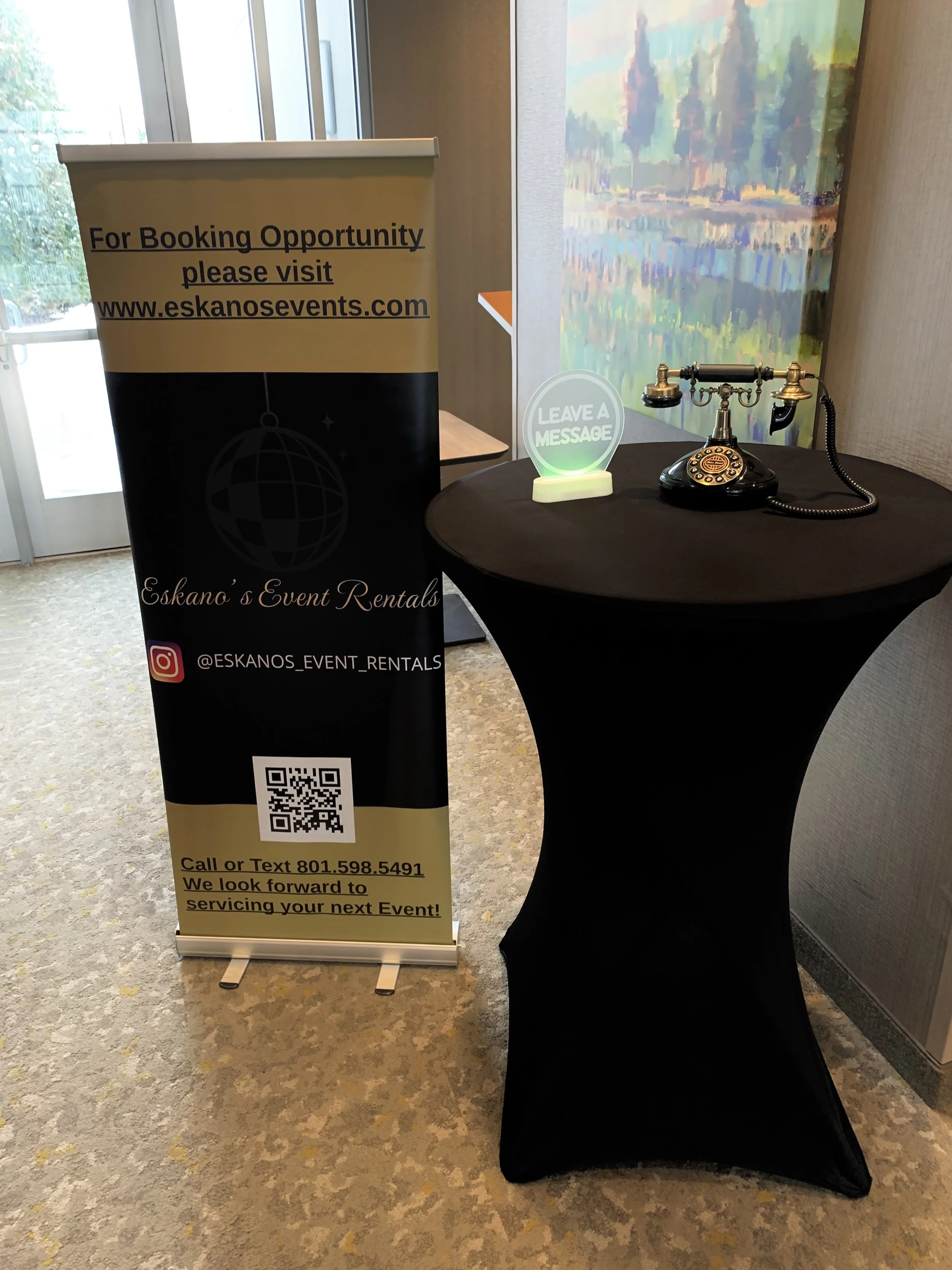 A standing yellow and black vertical banner with contact information and a QR code for Eskano's Event Rentals, positioned next to a round table with an antique telephone and a 'Leave a Message' sign in a room with a carpeted floor and waterfall artwo