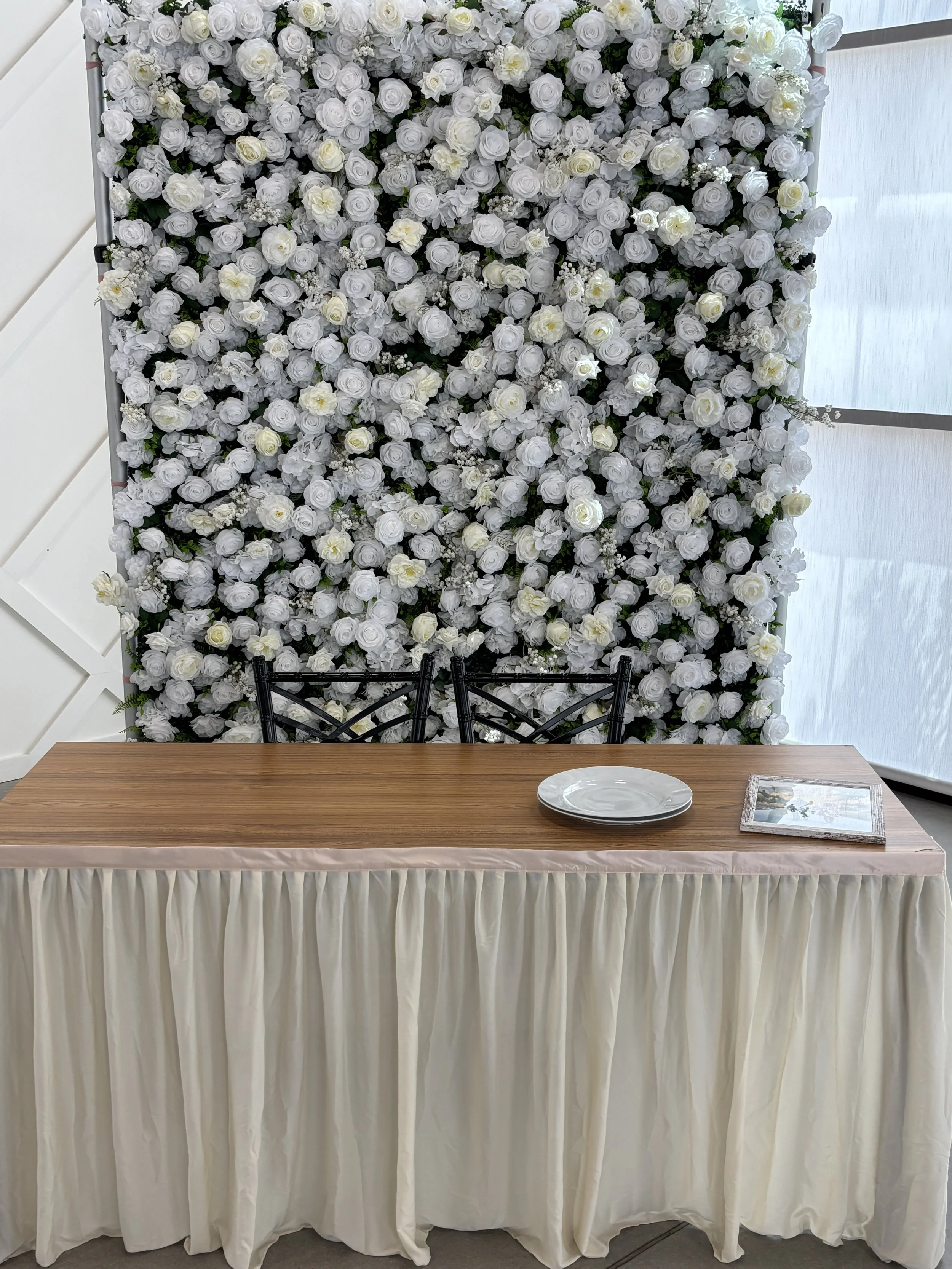 A table covered with a beige cloth, set with a white plate and a framed picture, in front of a backdrop of white roses and flowers, with black chairs and a sheer curtain on the right side