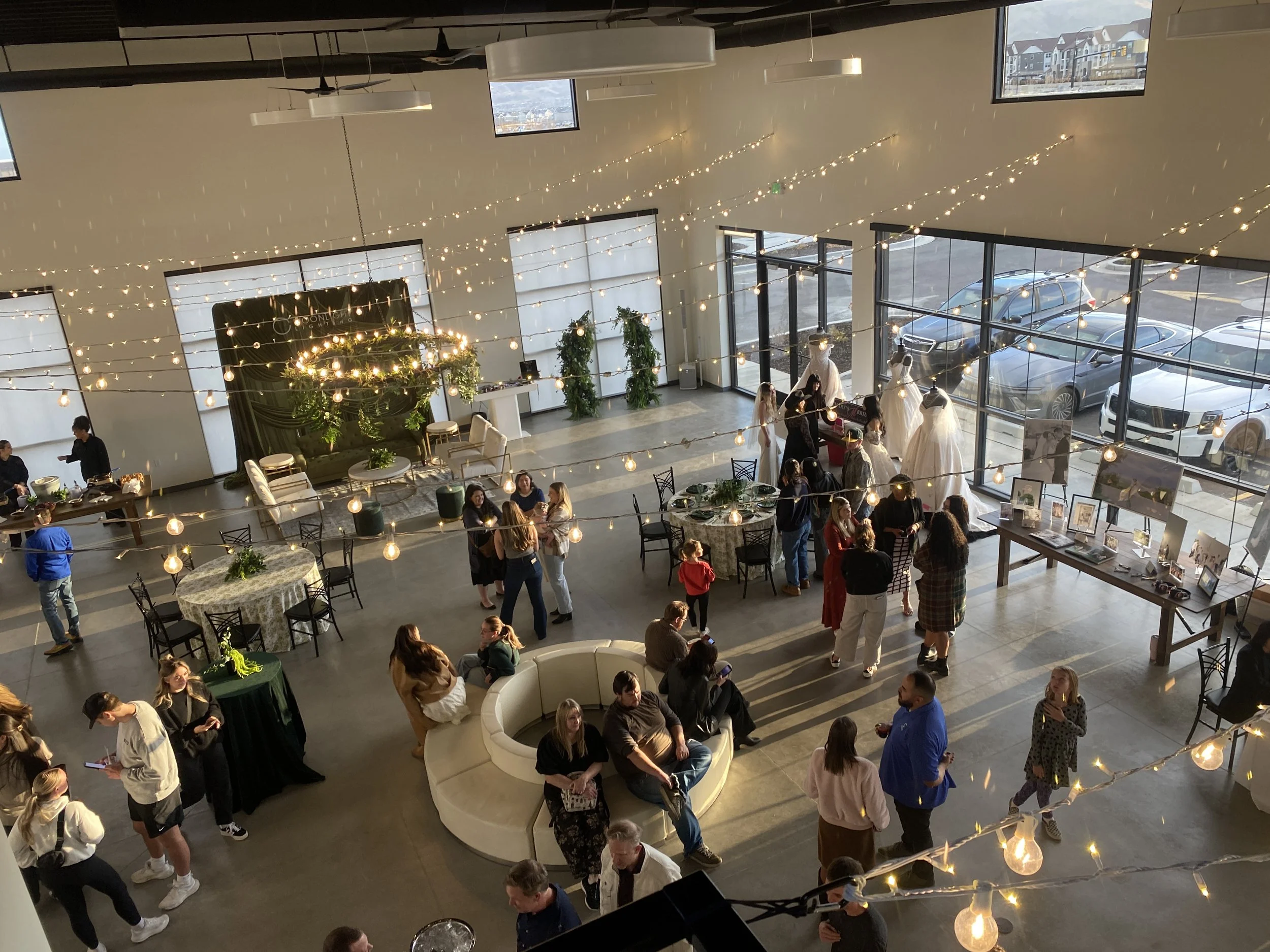 Indoor event space decorated with string lights, with people socializing, some standing and some sitting at round tables, and mannequins wearing dresses near large glass windows showing parked cars outside.