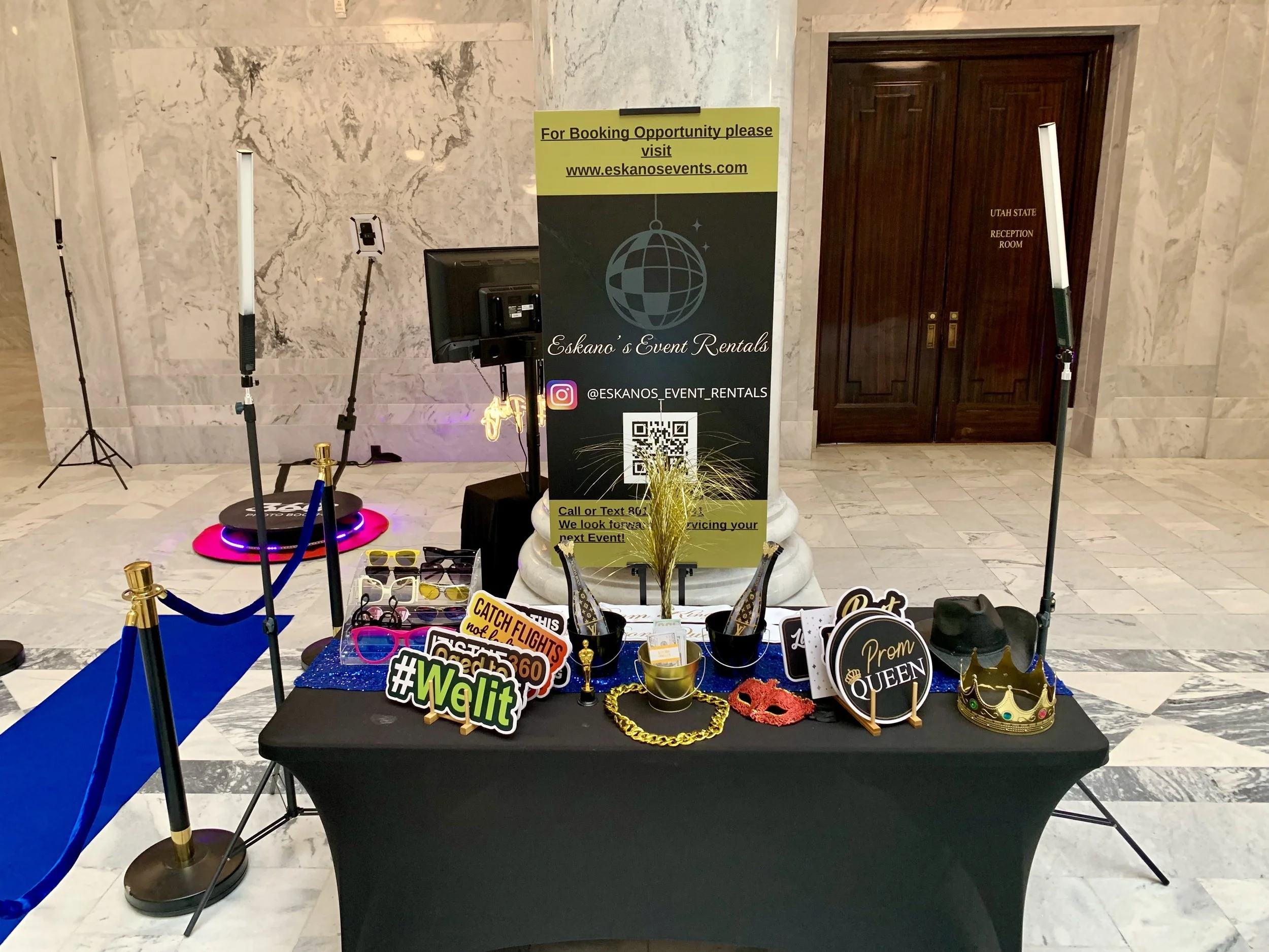 Table decorated with photo booth props, including tiaras, glasses, necklaces, and signs with words like 'Wild,' 'Celebrating,' and 'Queen.' Behind the table, there is a sign for Eskano's Event Rentals, with a globe logo and a QR code. The background includes a marble wall, a poster with booking information, and a door labeled 'Utah State Reception Room.' Equipment for lighting and photographs is set up around the table.