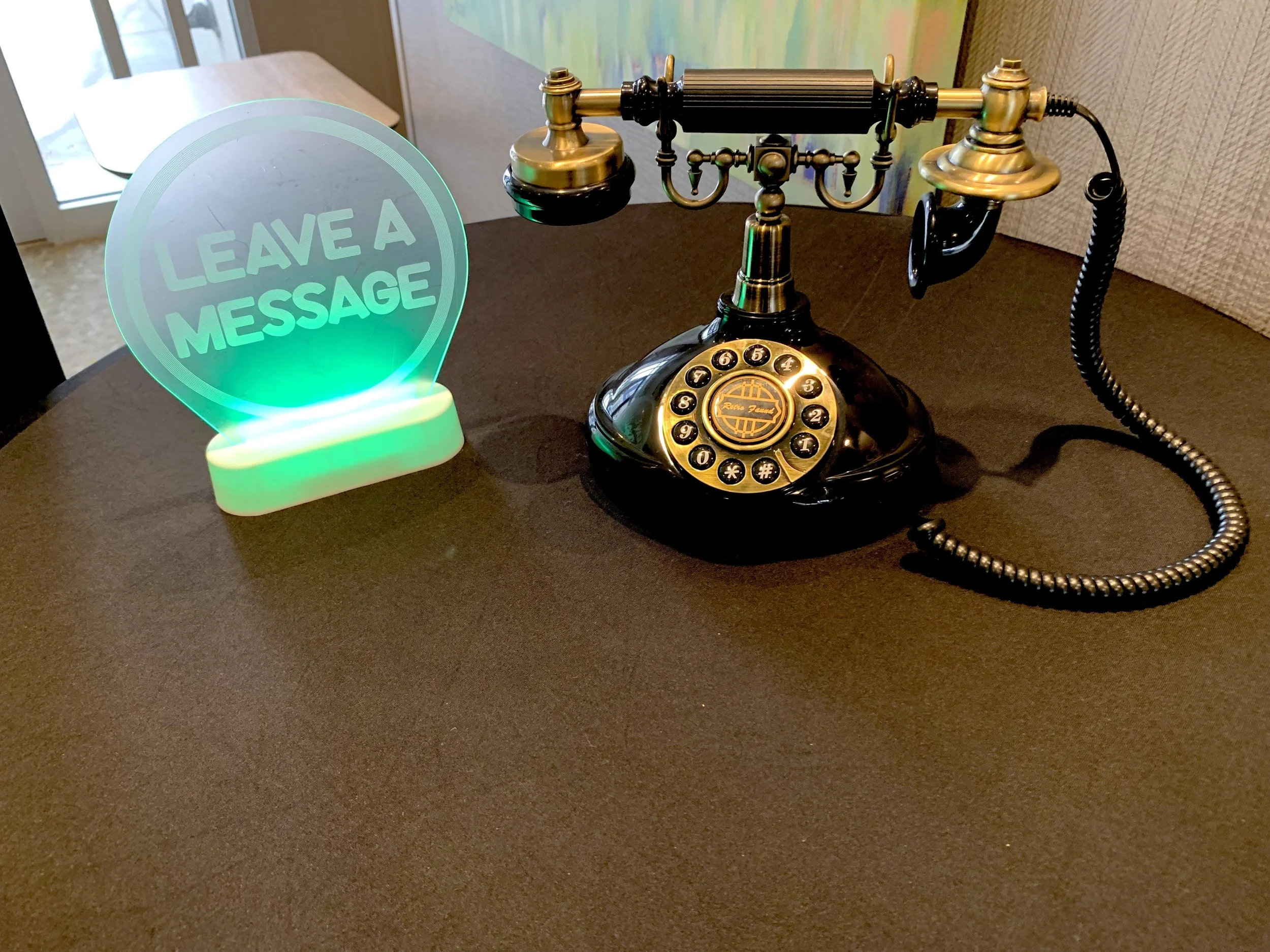 A vintage black and gold rotary dial telephone with a shiny finish, sitting on a dark table, next to a colorful illuminated sign that says "Leave a Message."