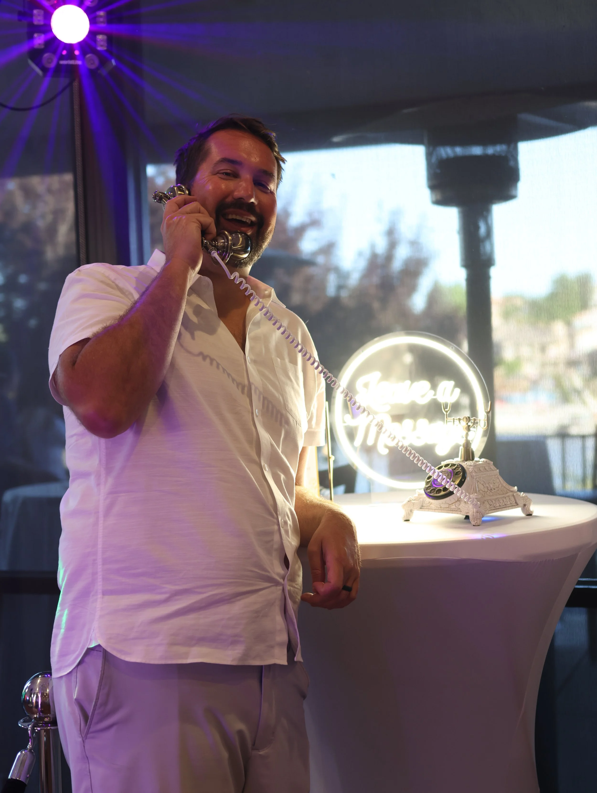 A man with a beard and wearing a light-colored shirt is smiling and talking on a rotary phone. Behind him, there's a lit circular sign with the name 'Fremont'. The setting appears to be a dimly lit venue with colorful lights and a large window showin
