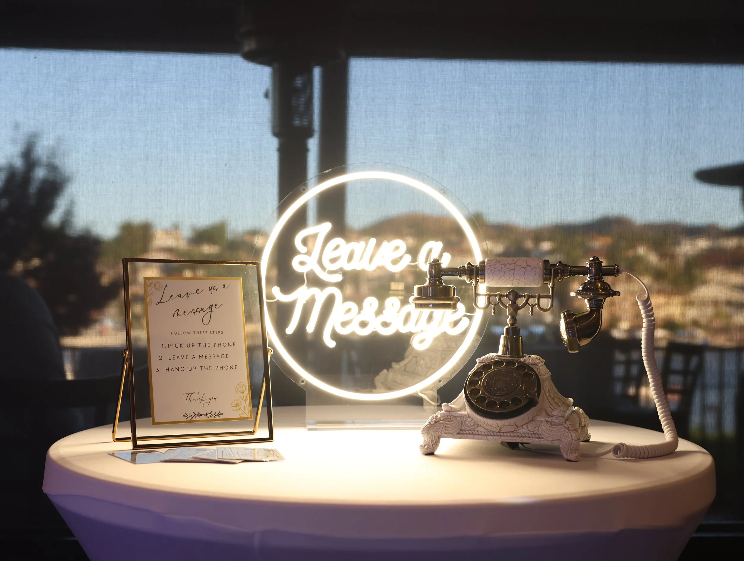 A vintage telephone with a rotary dial on a decorative stand, a lit neon sign that says 'Leave a message', and a framed note with step-by-step instructions for leaving a message, all on a round table with a window and a view of a sunset or sunrise sc