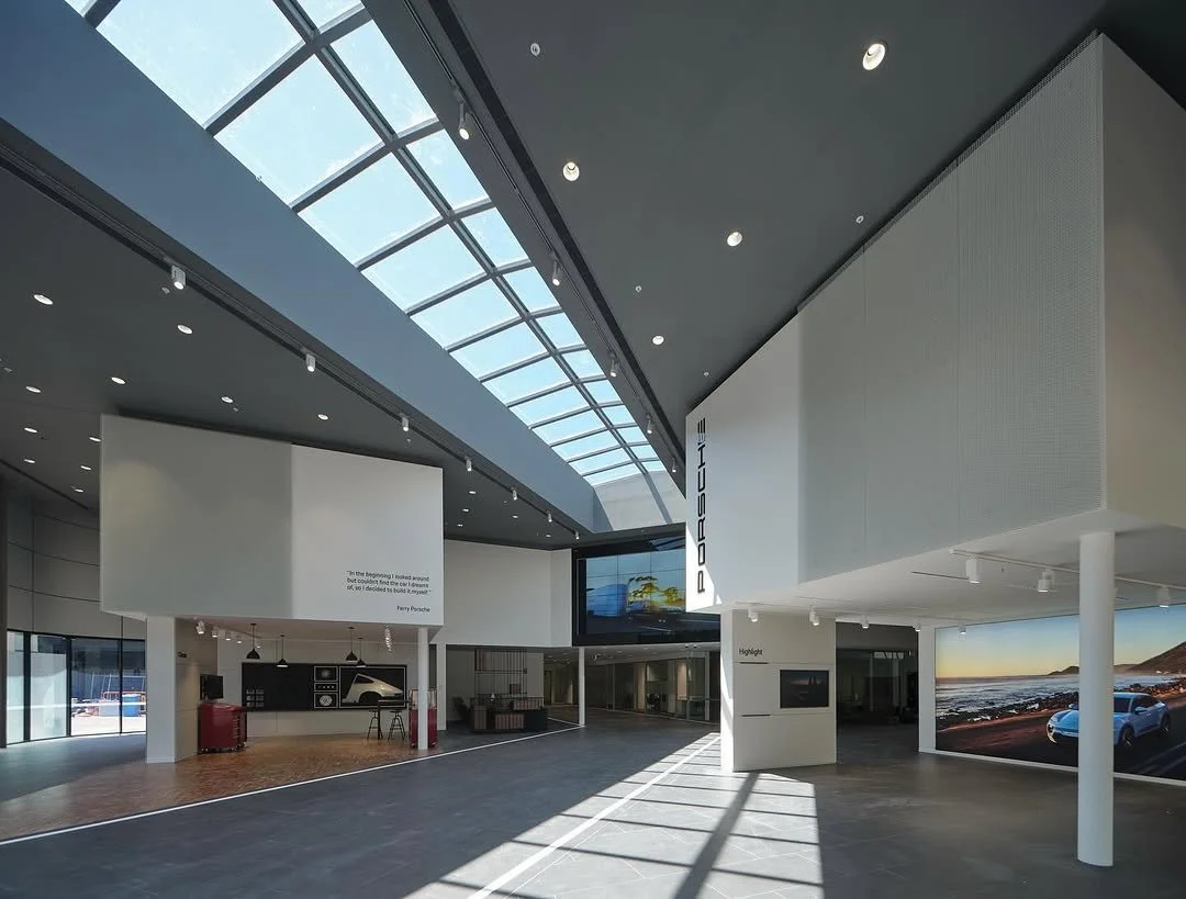 Interior of a modern Porsche dealership showroom with a glass ceiling, minimal decor, and a large digital screen displaying cars.