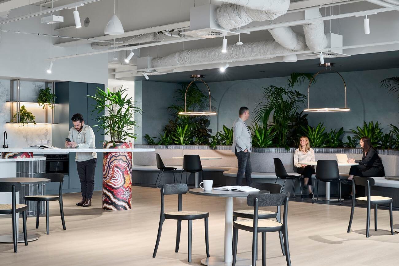 Modern office lounge space with people working and socializing, featuring plants, minimalist furniture, and contemporary lighting.