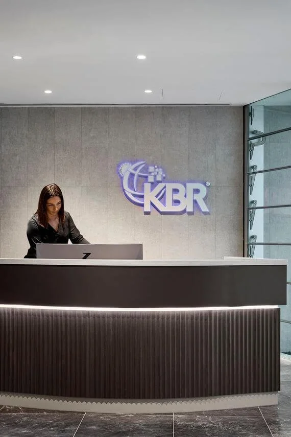 Reception desk with a woman working on a computer in front of a wall with a purple illuminated logo reading 'KBR'.