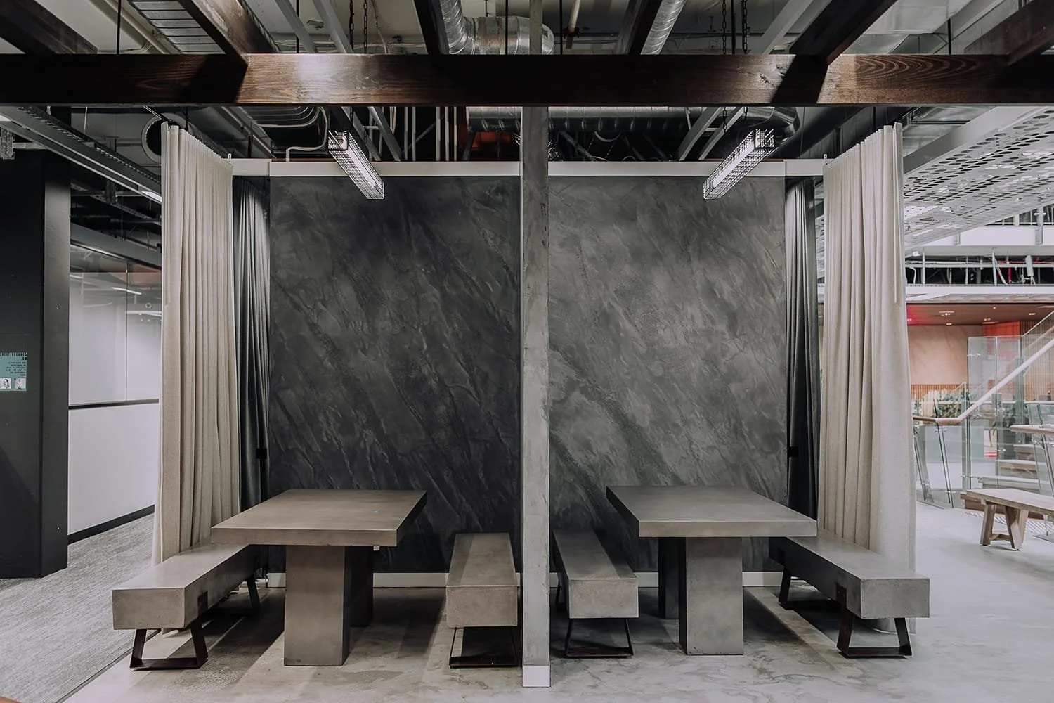 Modern conference or meeting space with two separate seating areas, divided by a concrete partition, featuring concrete tables and benches, with a dark textured wall backdrop and white curtains on either side.