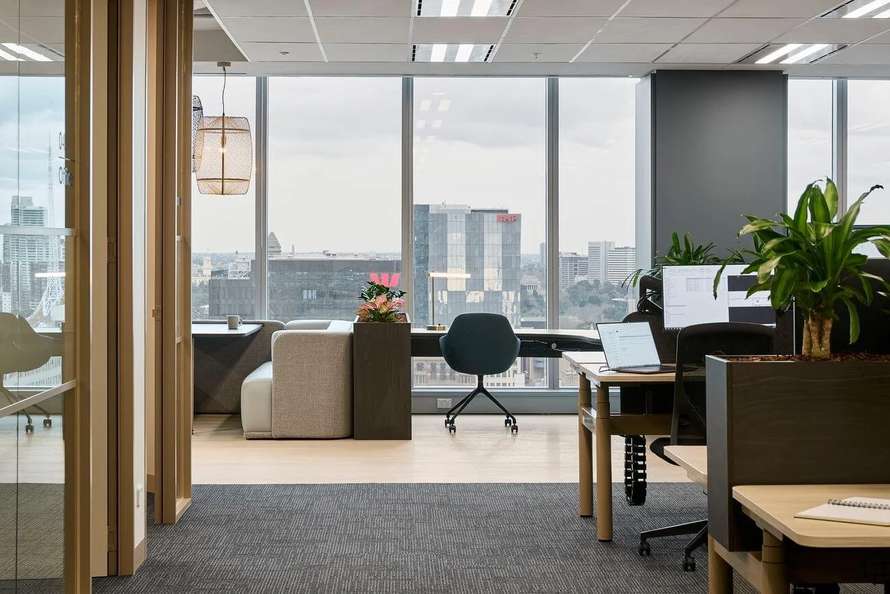 Modern office with large windows, desks with computers, chairs, computers showing spreadsheets, potted plants, a lounge area with sofa and a table, city view in the background.