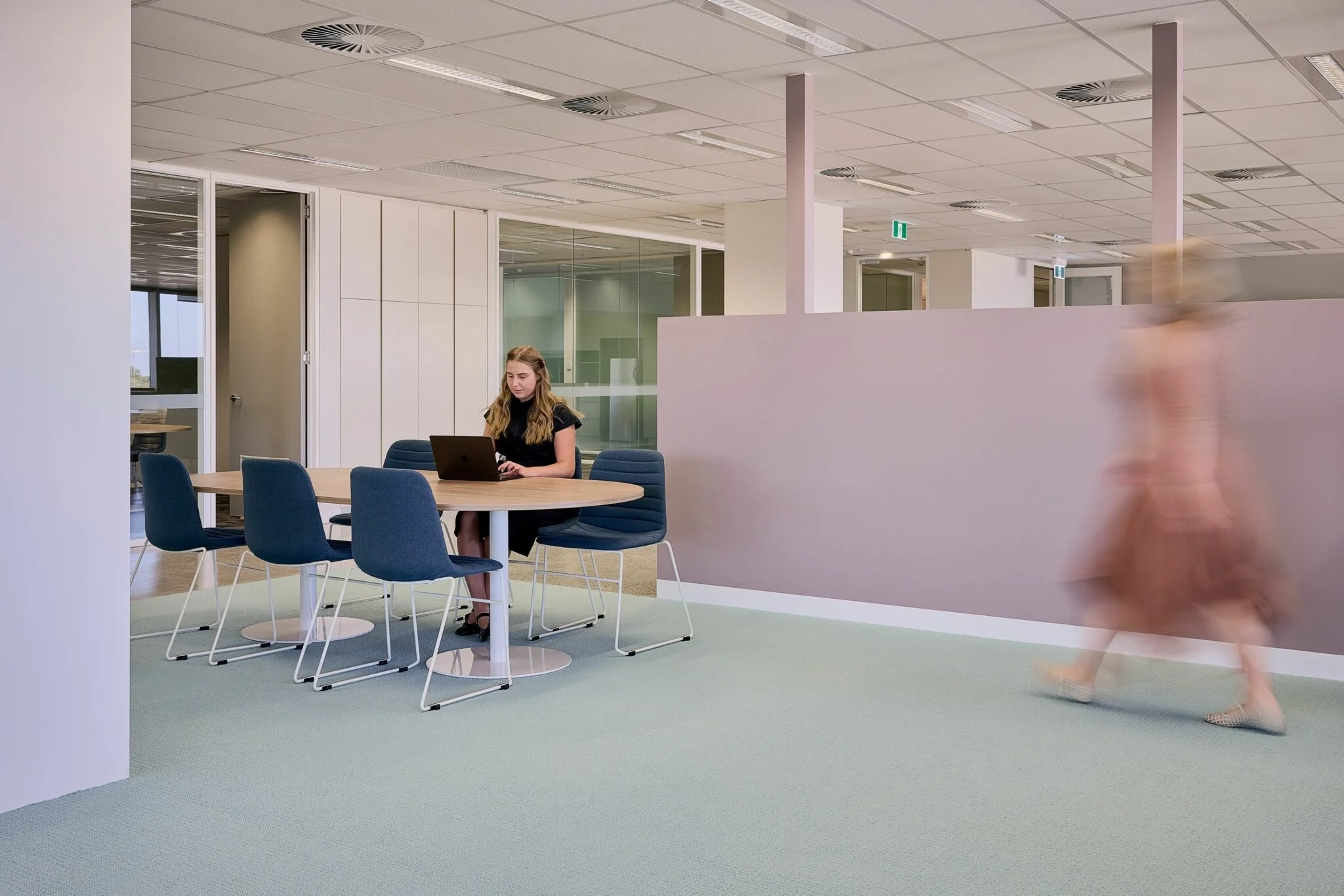 An office setting with a woman sitting at a table working on a laptop, and a blurred person walking past in the foreground. The office has modern decor with light-colored walls, a grid ceiling, and glass partition walls.