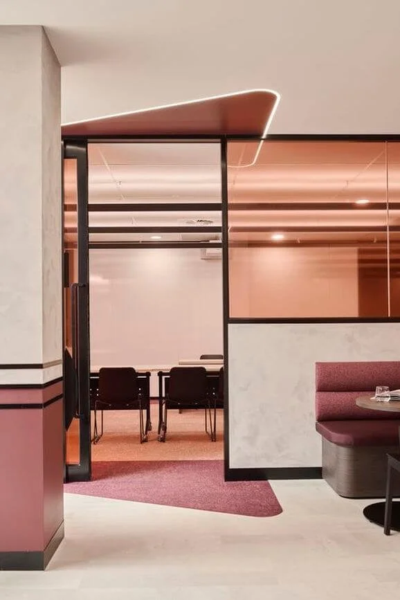 Modern interior with a glass partition, black framed sliding doors, a black table with chairs in a bright room, and a pink booth seating area with a glass on the table.