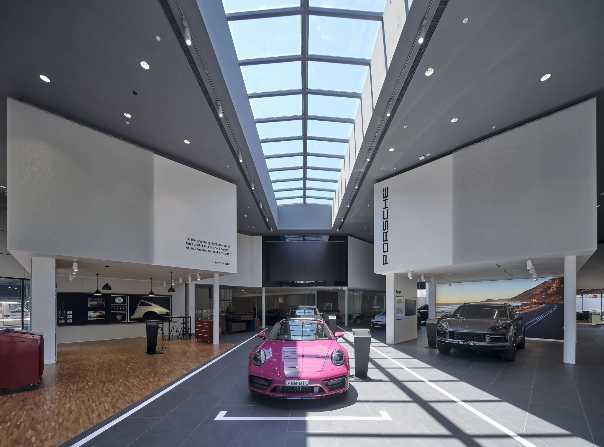 Inside a Porsche dealership showroom with a pink sports car in the foreground, black and silver cars in the background, and a large skylight ceiling letting in natural light.