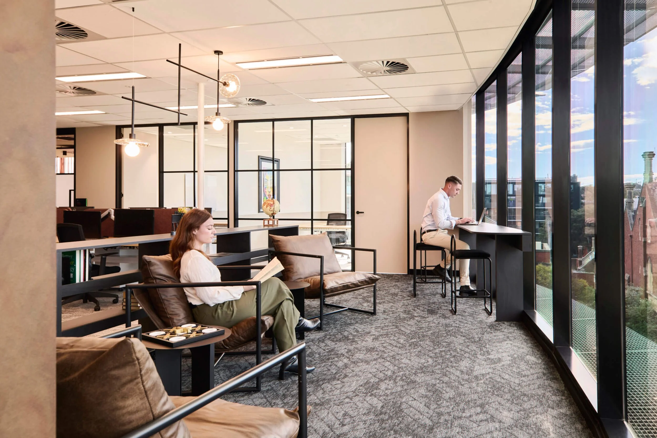 A modern office space with large window walls showing a city view, two people working on laptops, a woman sitting on a brown leather couch reading, and a man sitting at a high table using his laptop, with pendant lights hanging from the ceiling.