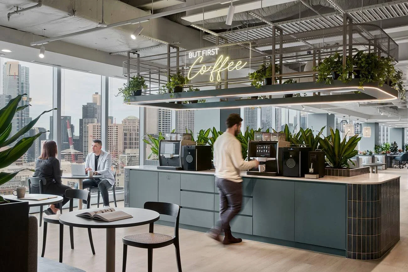 Modern office coffee station with a neon sign saying 'But First Coffee', large windows with city skyline views, and people enjoying coffee and conversation.