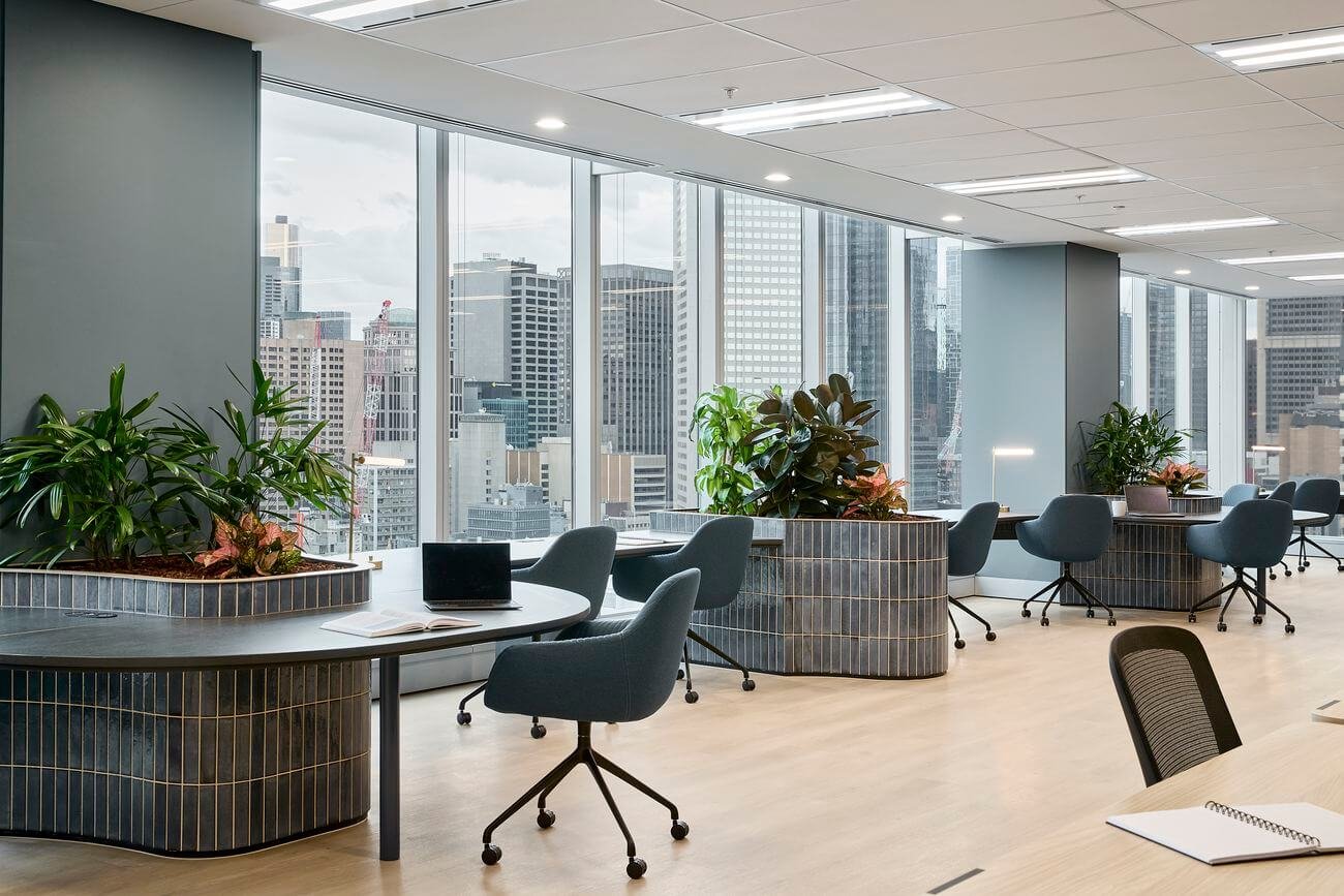 Modern office space with large windows, city skyline view, gray chairs, circular desks with potted plants, and notebooks on the table.
