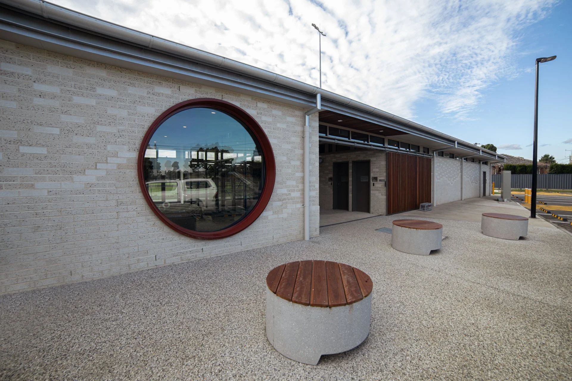 Exterior view of a modern building with a large circular window, light-colored brick walls, and outdoor seating with round concrete tables and wooden tops. There is a parking lot with yellow-striped spaces and street lamps under a partly cloudy sky.