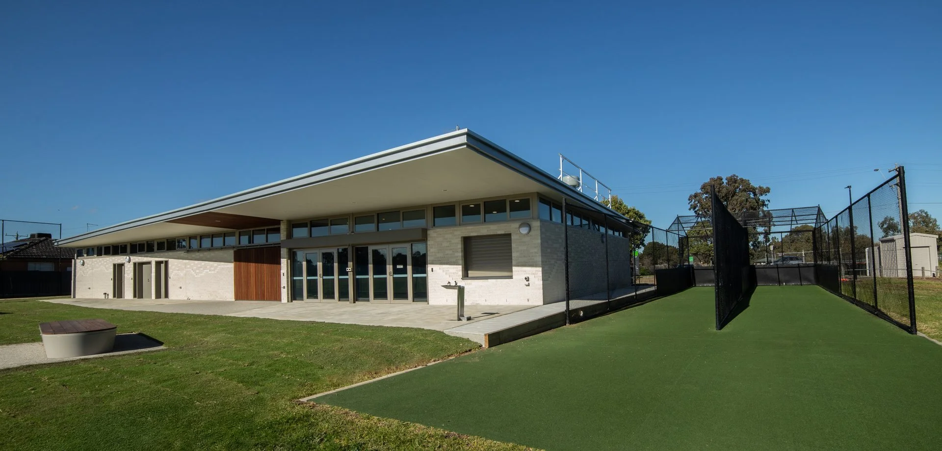 Modern building with large windows and a flat roof next to a fenced tennis court and green lawn, under a clear blue sky.
